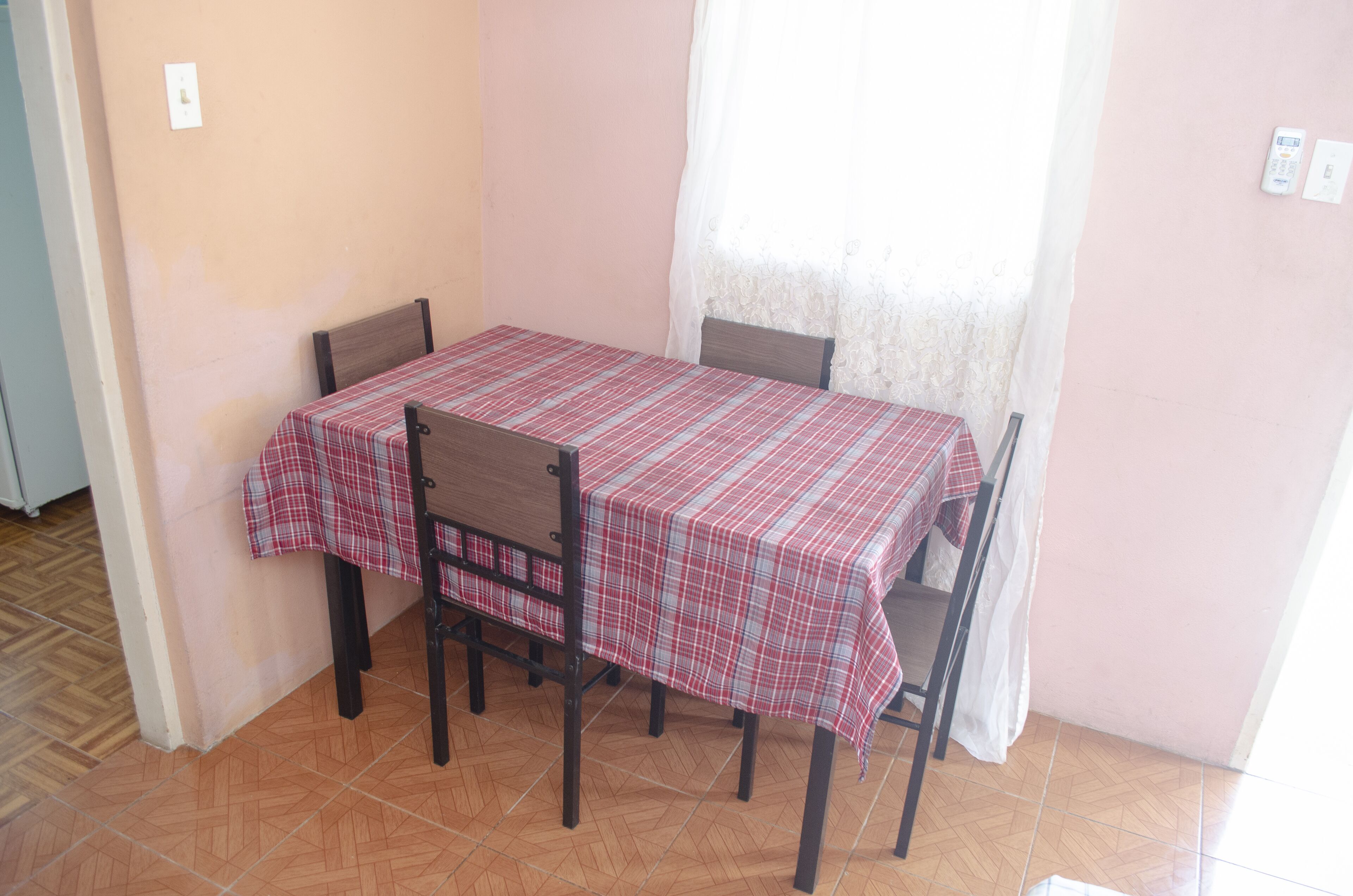 Un coin repas avec une table recouverte d’une nappe à carreaux, quatre chaises et une fenêtre ornée d’un rideau léger.
