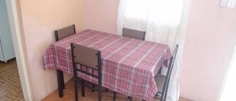 Un coin repas avec une table recouverte d’une nappe à carreaux, quatre chaises et une fenêtre ornée d’un rideau léger.