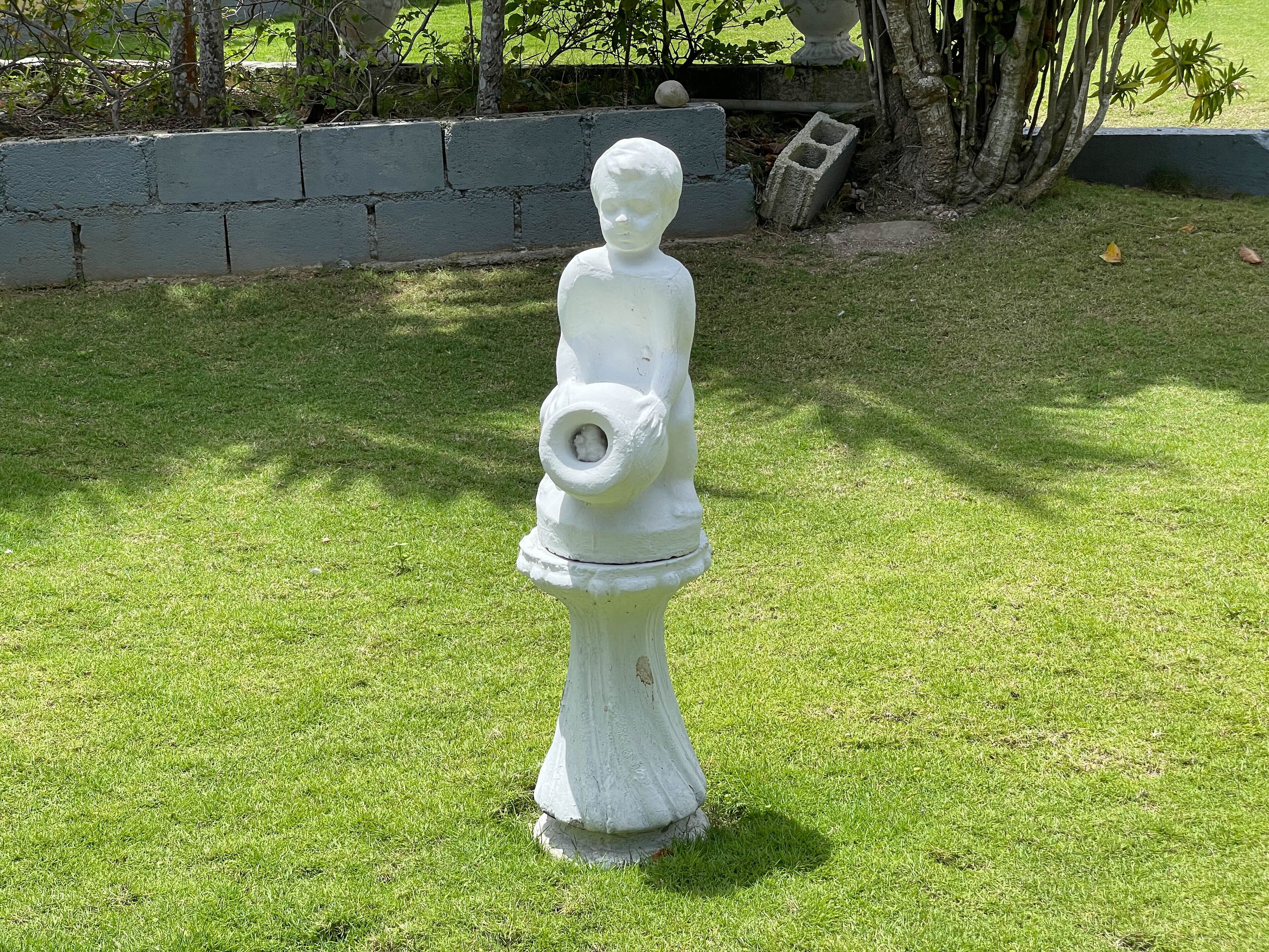Una estatua blanca de un niño sosteniendo una manguera en un área con césped, con un muro de piedra y árboles al fondo.