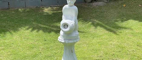 A white statue of a child holding a hose in a grassy area with a stone wall and trees in the background.