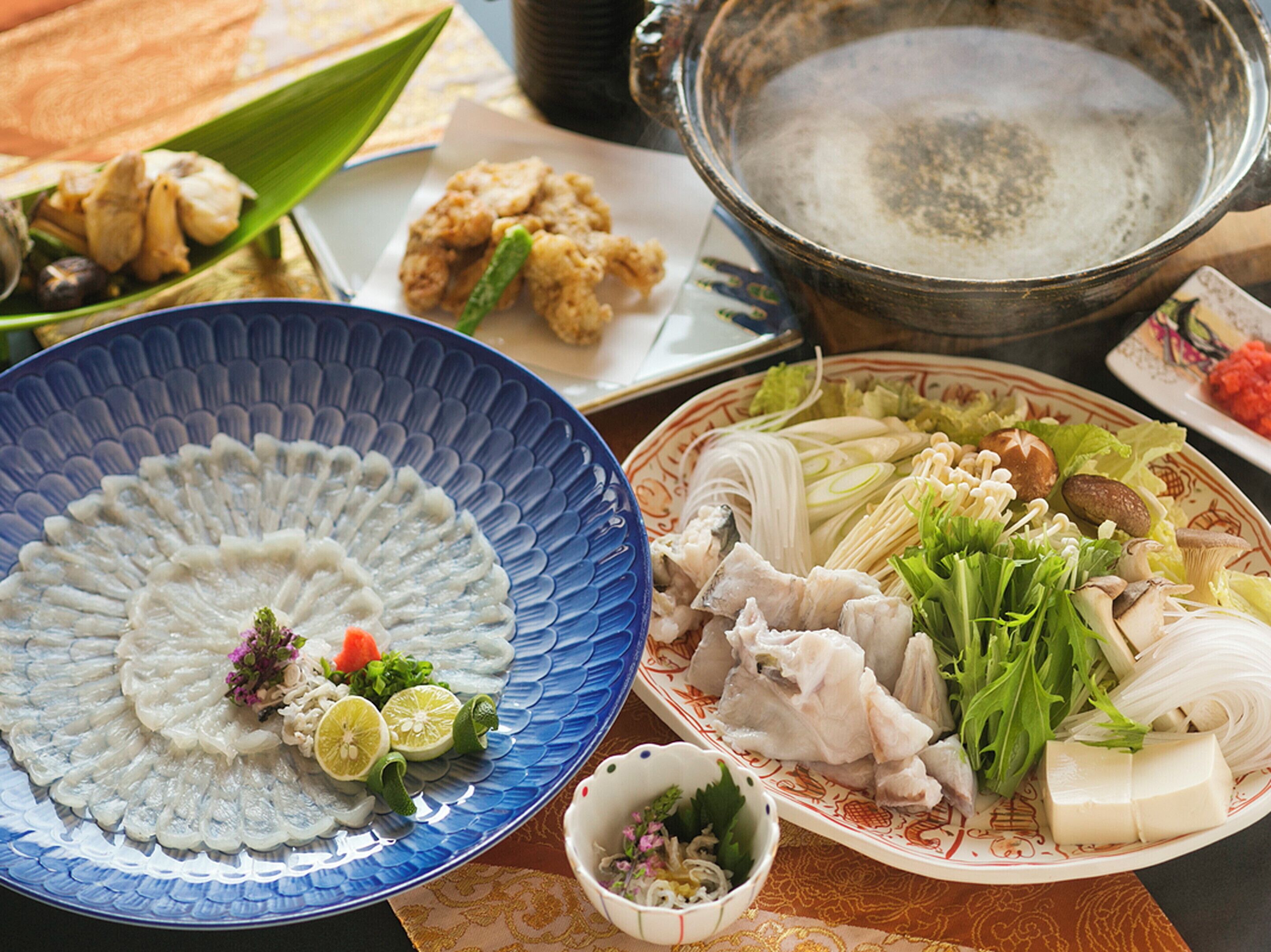 A variety of dishes including a blue plate with sashimi, a plate with vegetables and tofu, and a pot of boiling water.