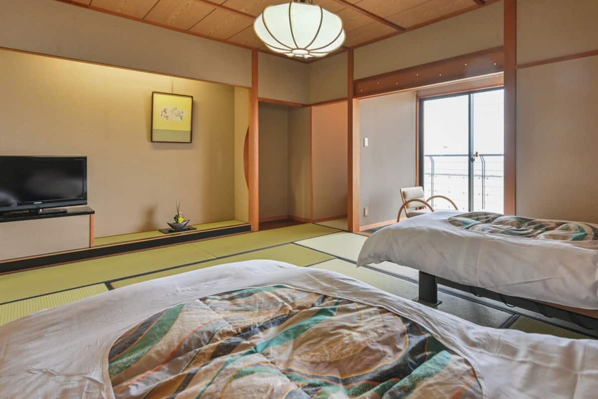 A traditional Japanese room with tatami flooring, two beds, a television, and a sliding door leading to a balcony.