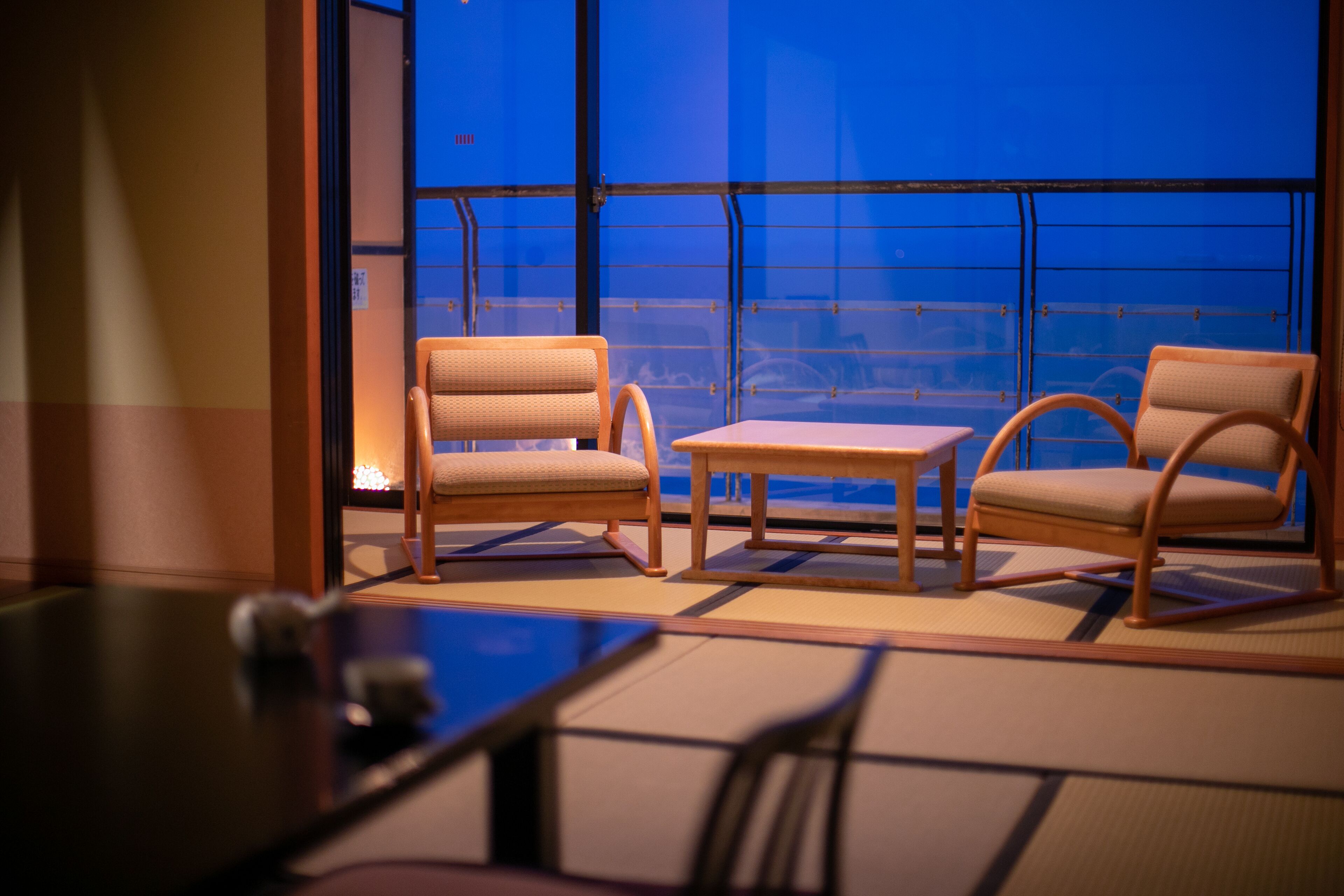 A room with tatami flooring, two wooden chairs, a small table, and a view through large windows.