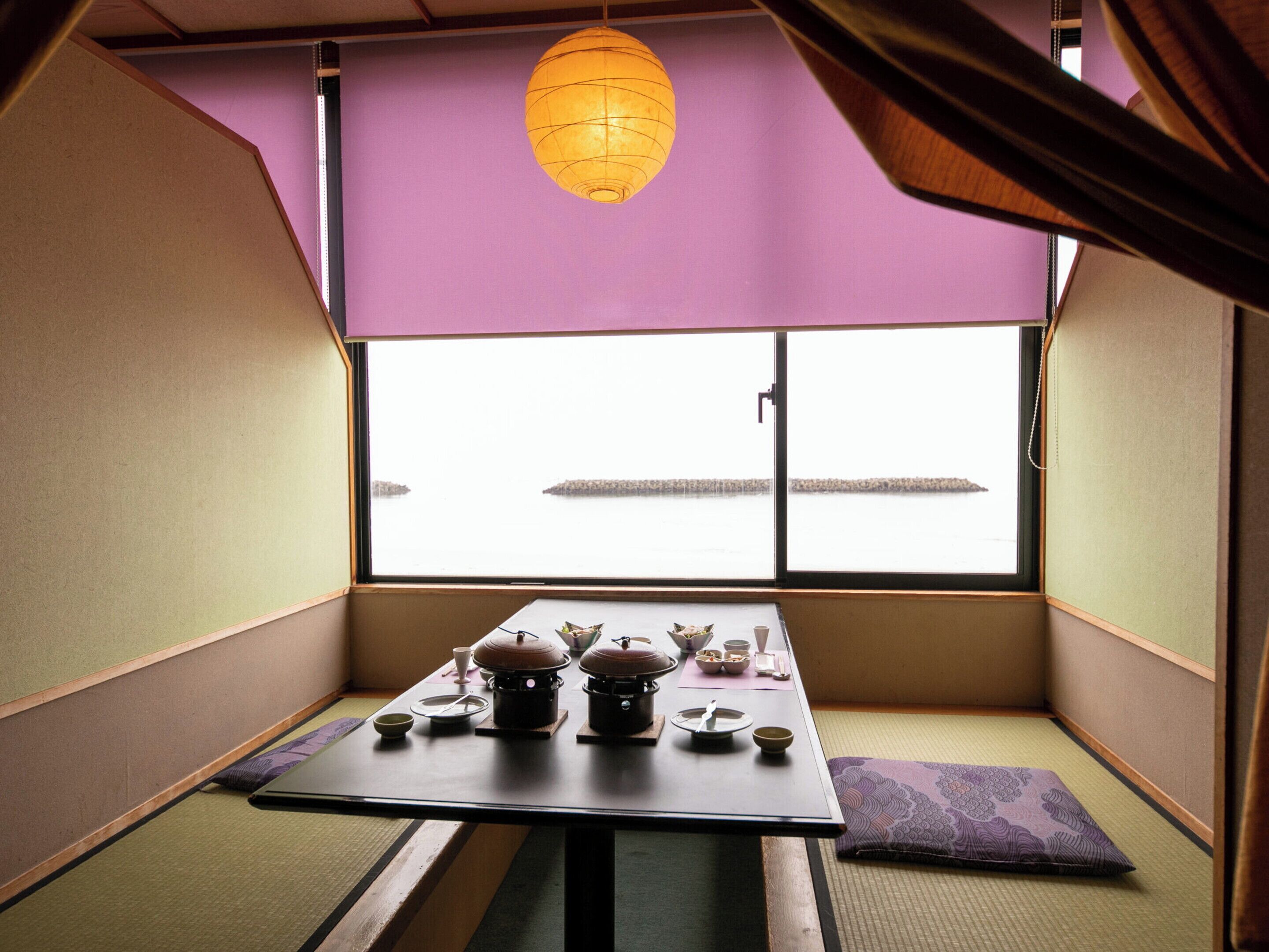 A traditional Japanese room with tatami flooring, a low table with a tea set, and a view through a large window.