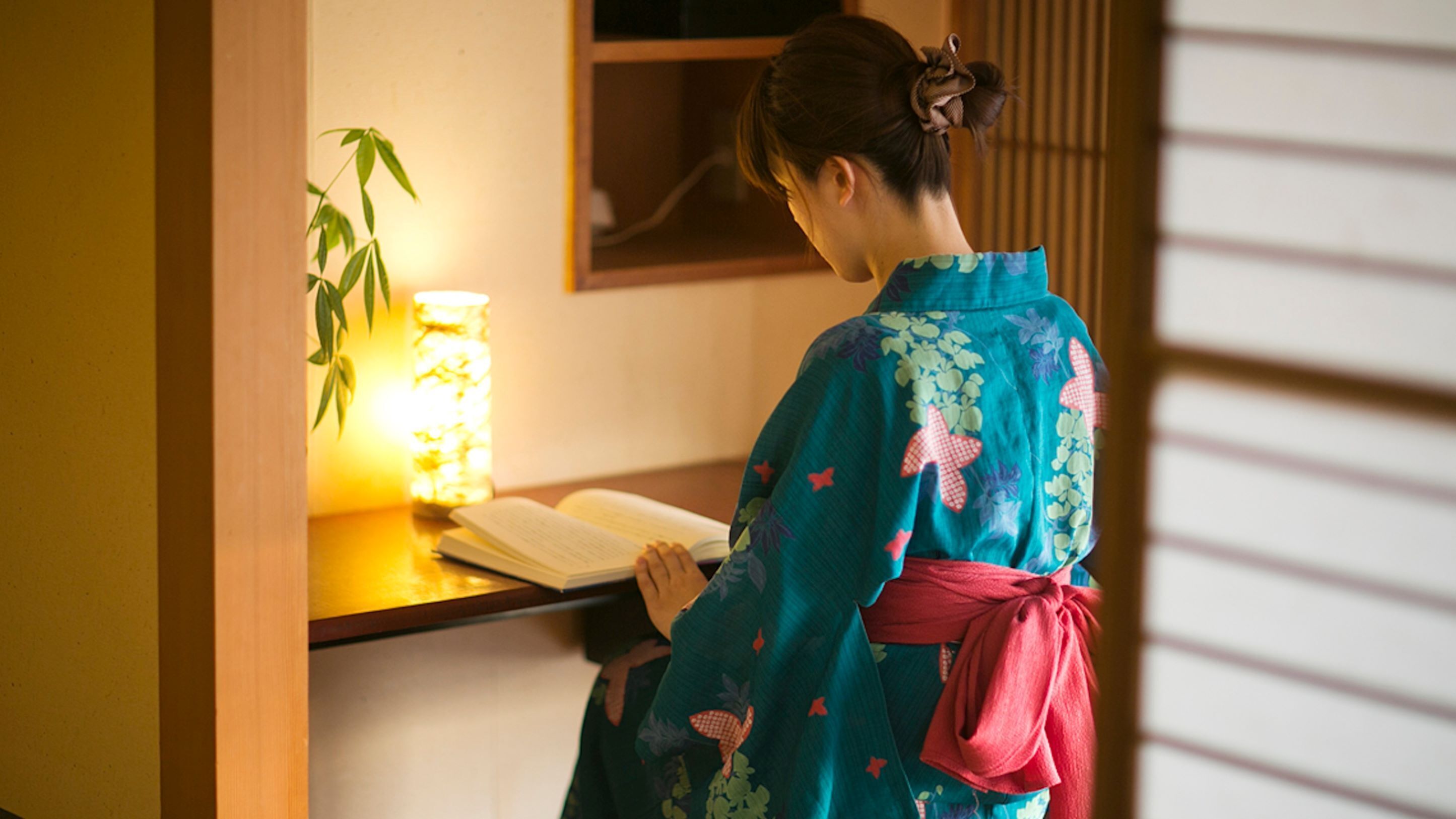 Una persona vestida con traje tradicional leyendo un libro en una habitación con un estante de madera, una vela encendida y una planta.