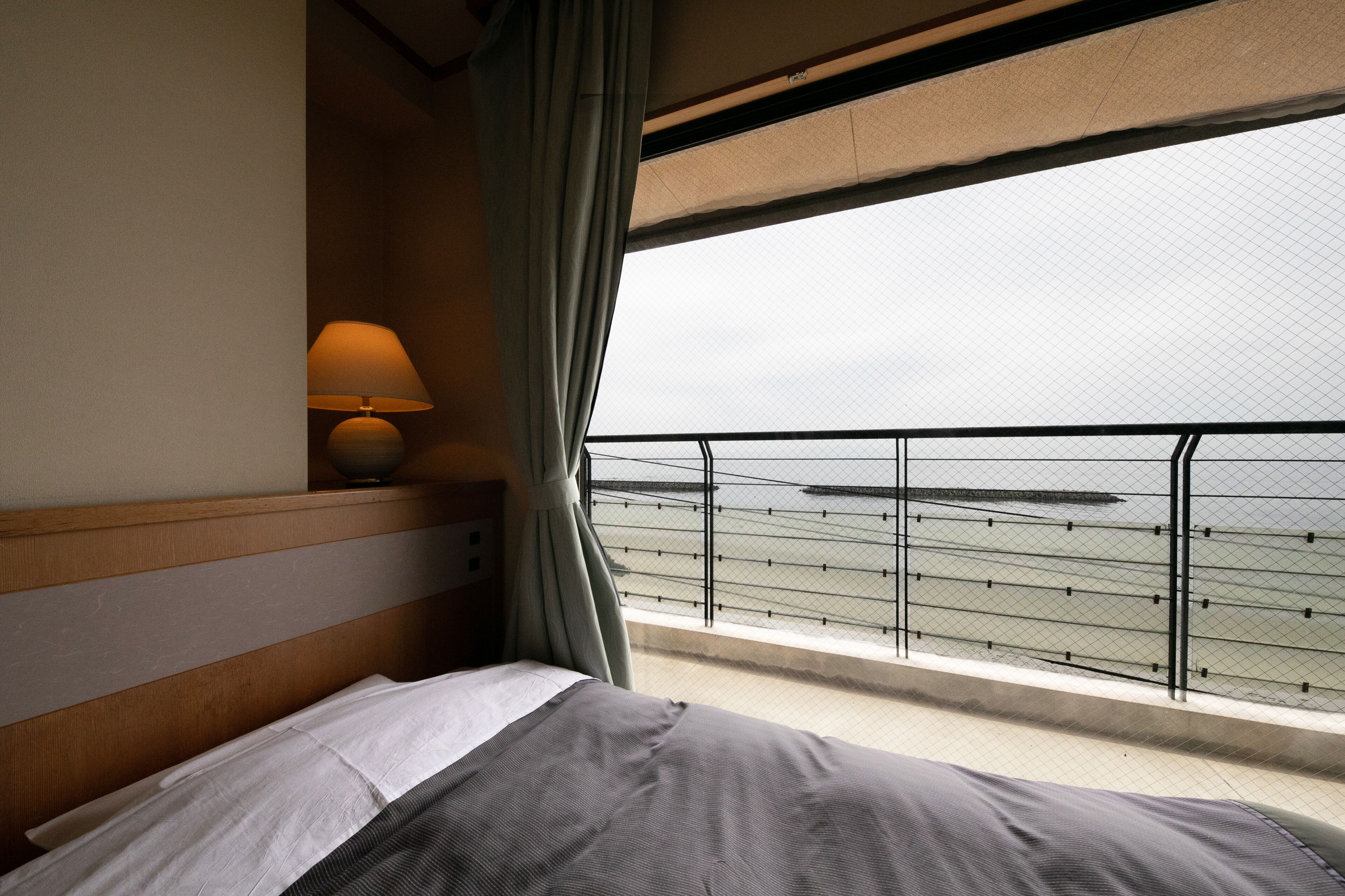 A hotel room with a bed, a lamp, and a view of the sea through a balcony with a railing.