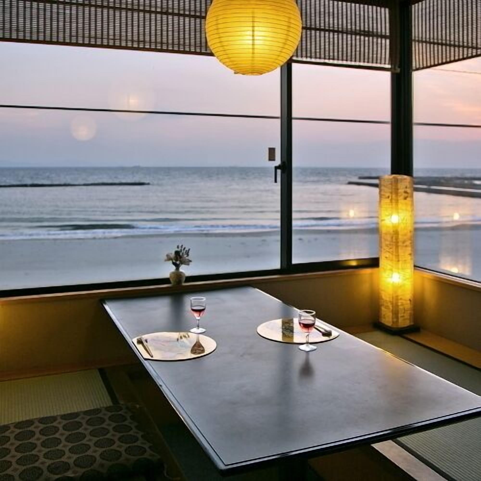 A room with a view of the ocean, a table with two glasses of wine, a vase with flowers, and a traditional lantern.