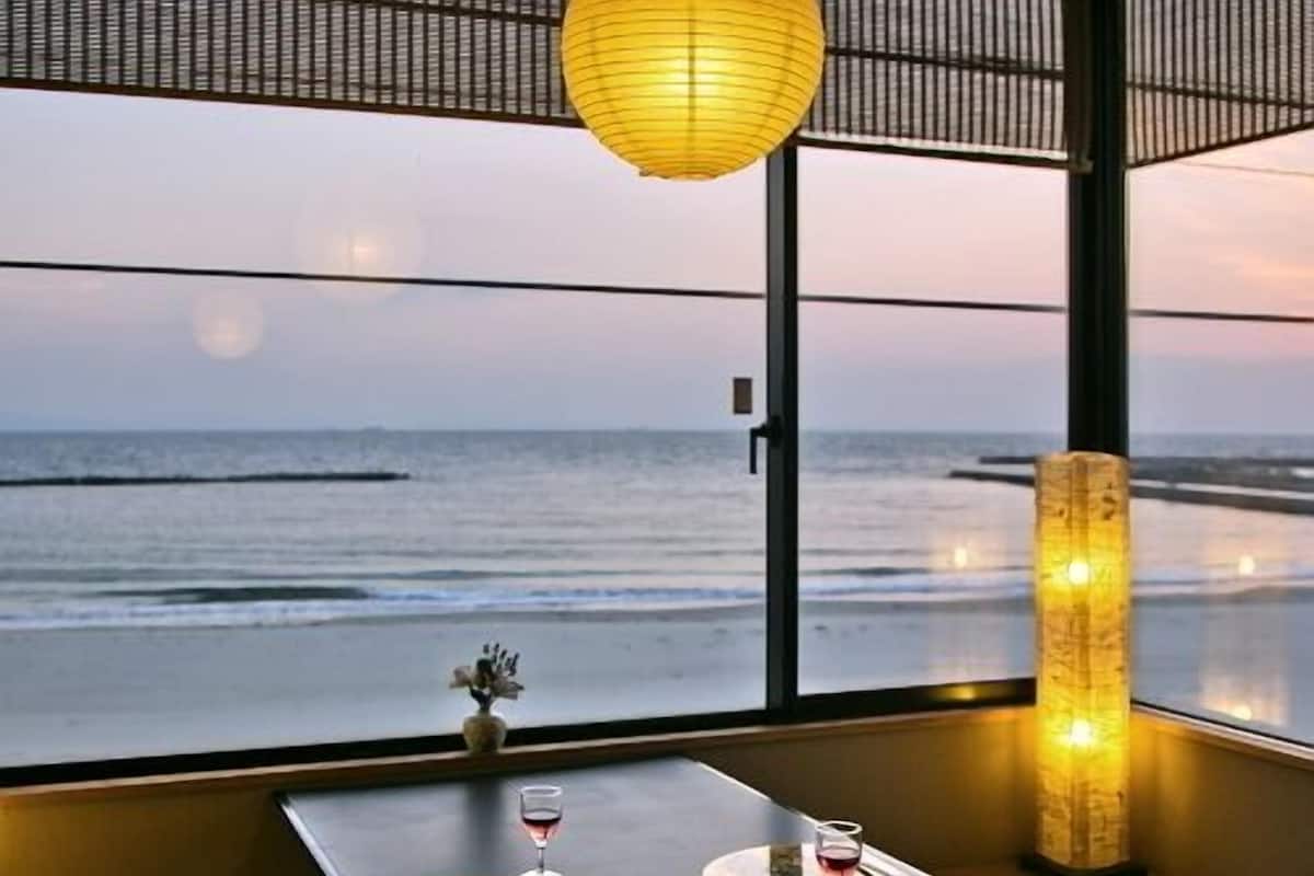 A room with a view of the ocean, a table with two glasses of wine, a vase with flowers, and a traditional lantern.