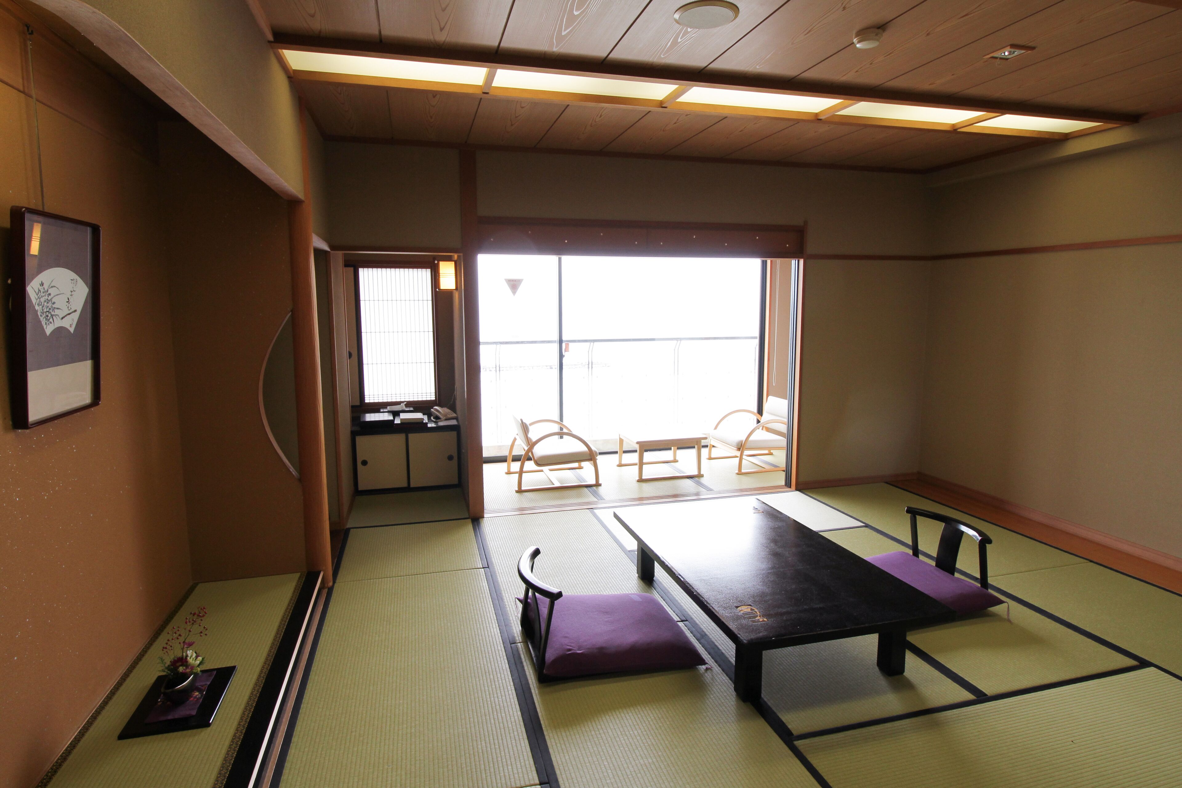Una habitación con esteras de tatami, una mesa baja y sillas, una pequeña mesa con un florero, y una puerta corredera que da a otra habitación.