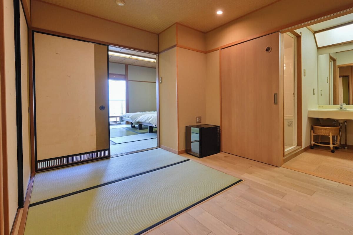 A traditional Japanese room with tatami flooring, a bed, a small cabinet, and a bathroom with a sink and mirror.