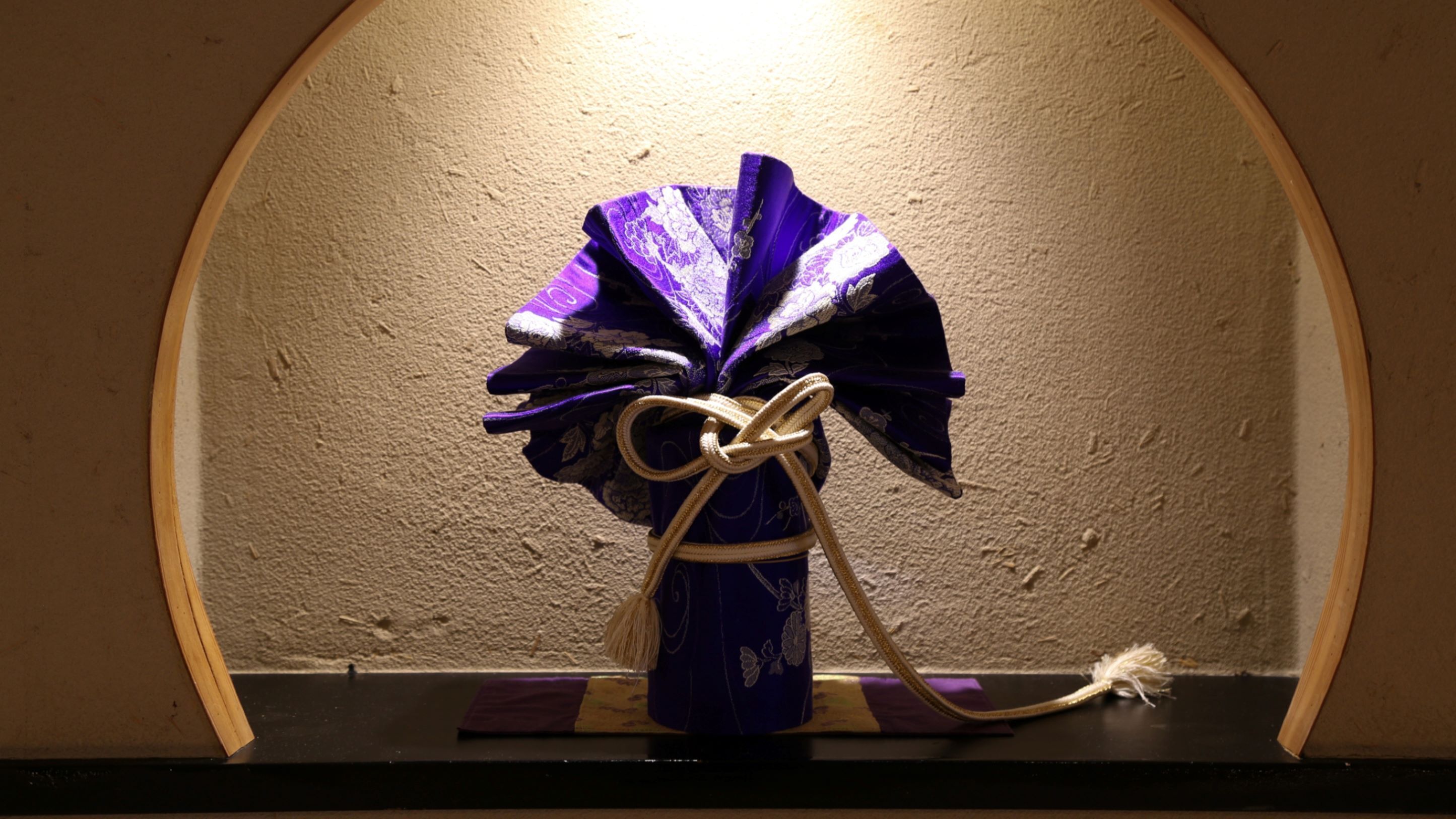A vase with a purple flower arrangement tied with a white string, placed on a black surface with a textured wall in the background.