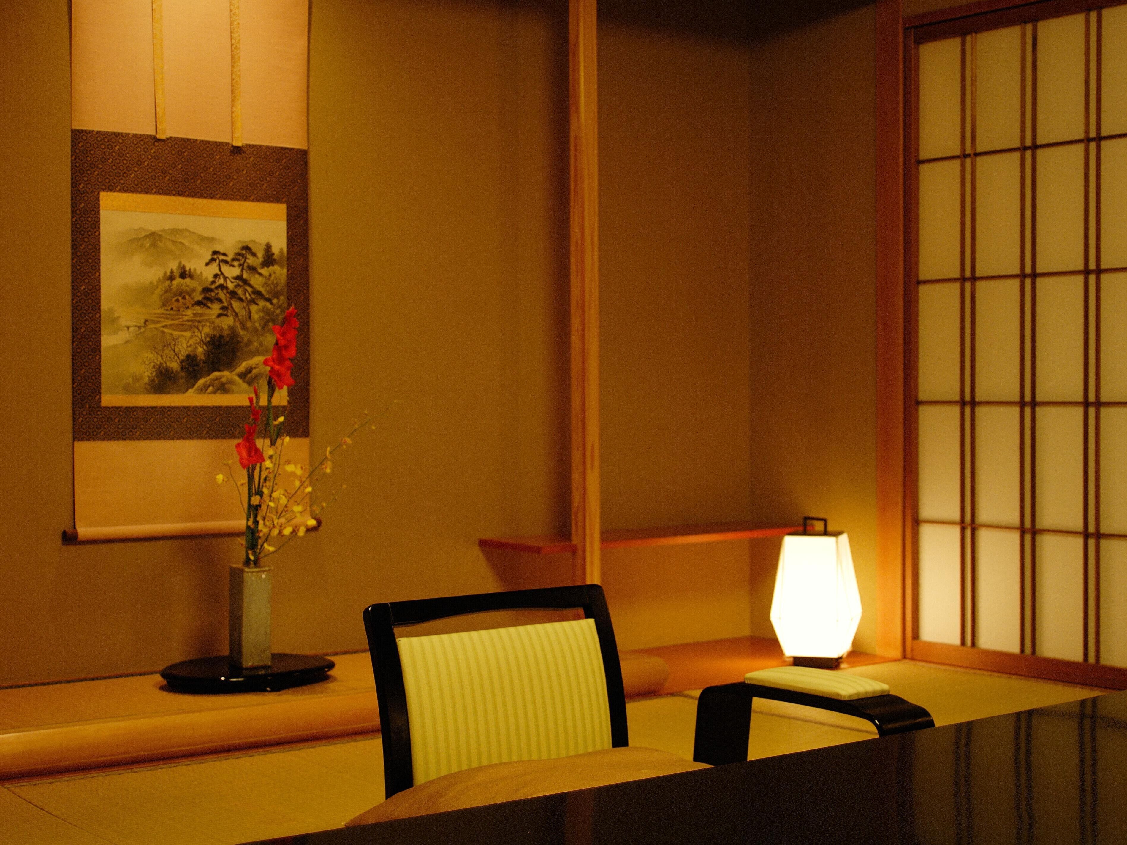 A traditional Japanese room with tatami flooring, a low table, a chair, a vase with flowers, a framed picture on the wall, and a shoji screen.