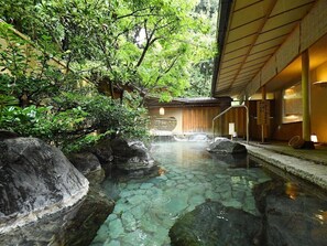 Un onsen japonais traditionnel, avec une source d’eau chaude claire, entouré de rochers et d’une végétation luxuriante.