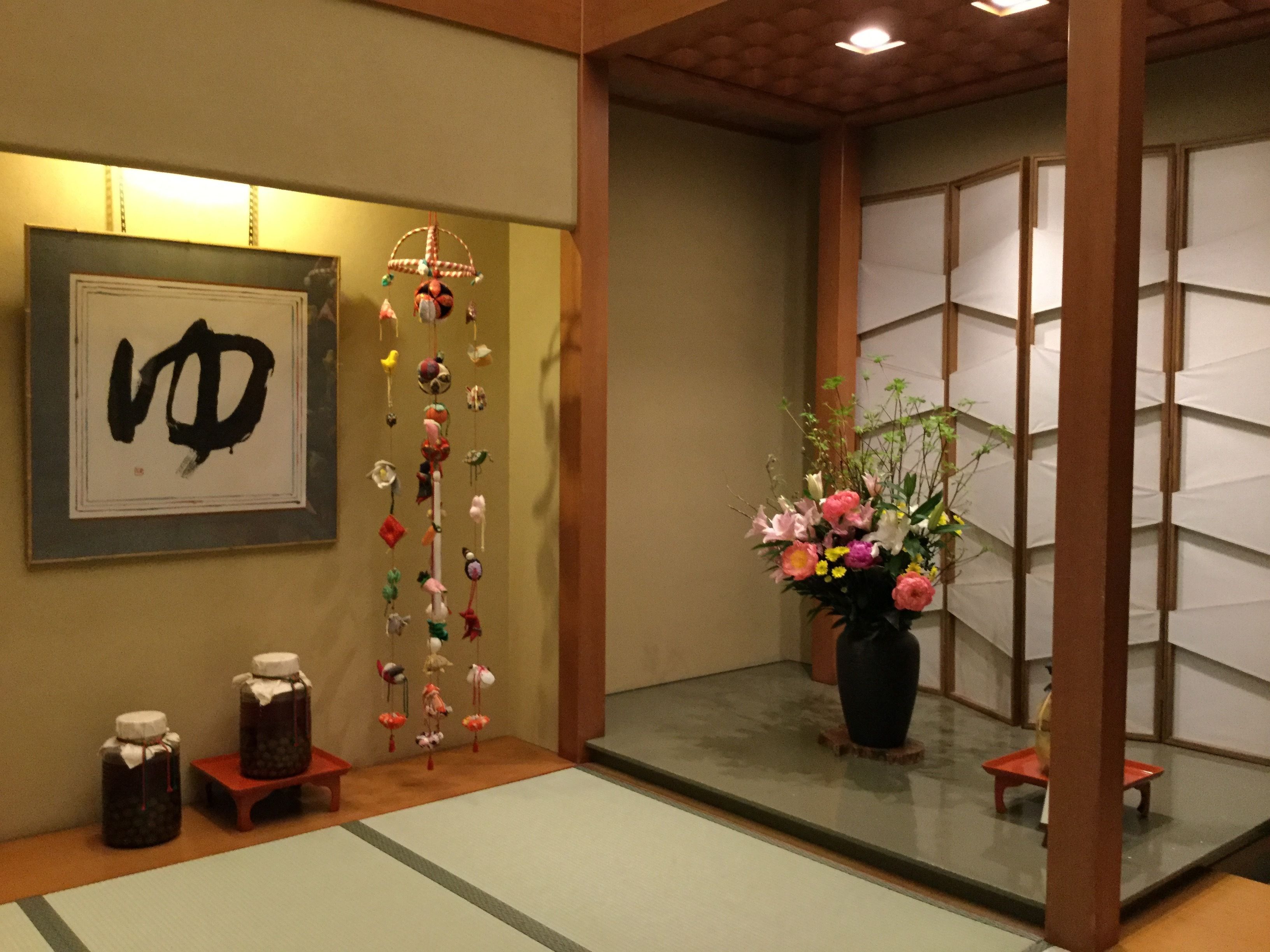 A traditional Japanese room with tatami flooring, a shoji screen, a framed calligraphy piece, a hanging ornament, a vase with flowers, and two sake bottles on a low table.