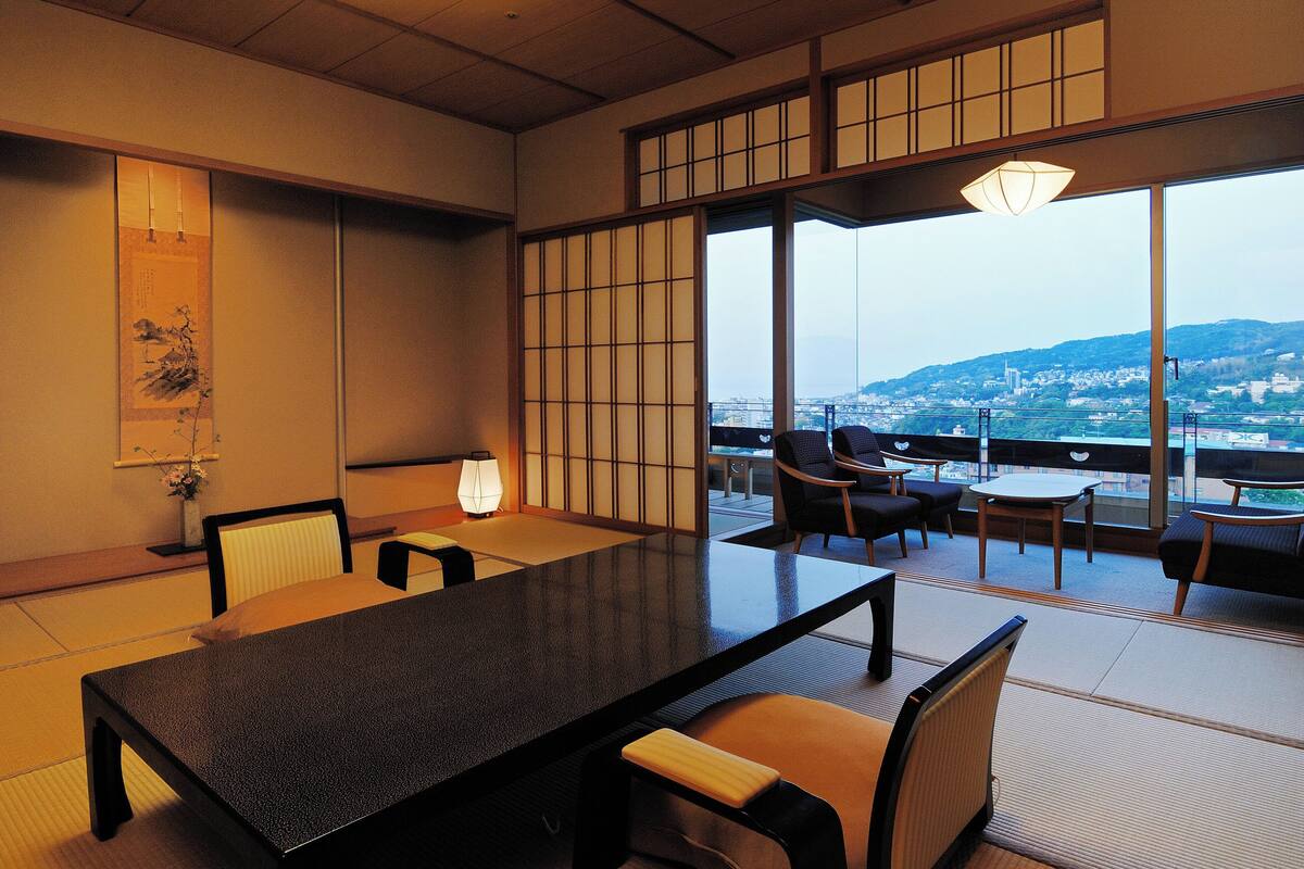A traditional Japanese room with tatami flooring, a low table, and chairs. There is a large window with a view of a cityscape.