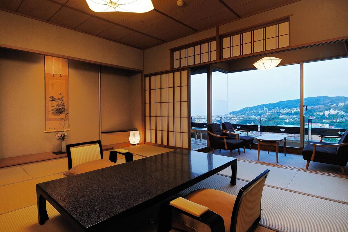 A traditional Japanese room with tatami flooring, a low table, and chairs. It features large windows with a view of a cityscape.