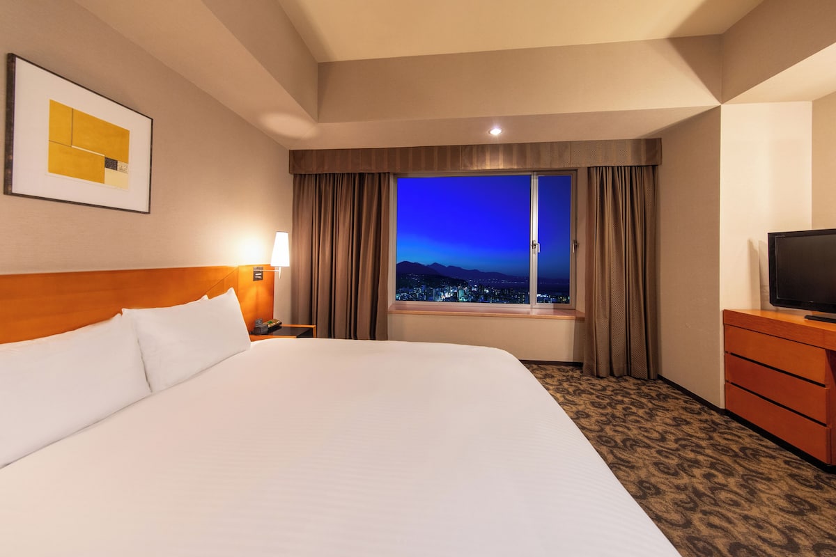 Habitació d'hotel amb un llit gran, televisió, tauleta de nit, làmpada i finestra amb vista a la ciutat al capvespre.