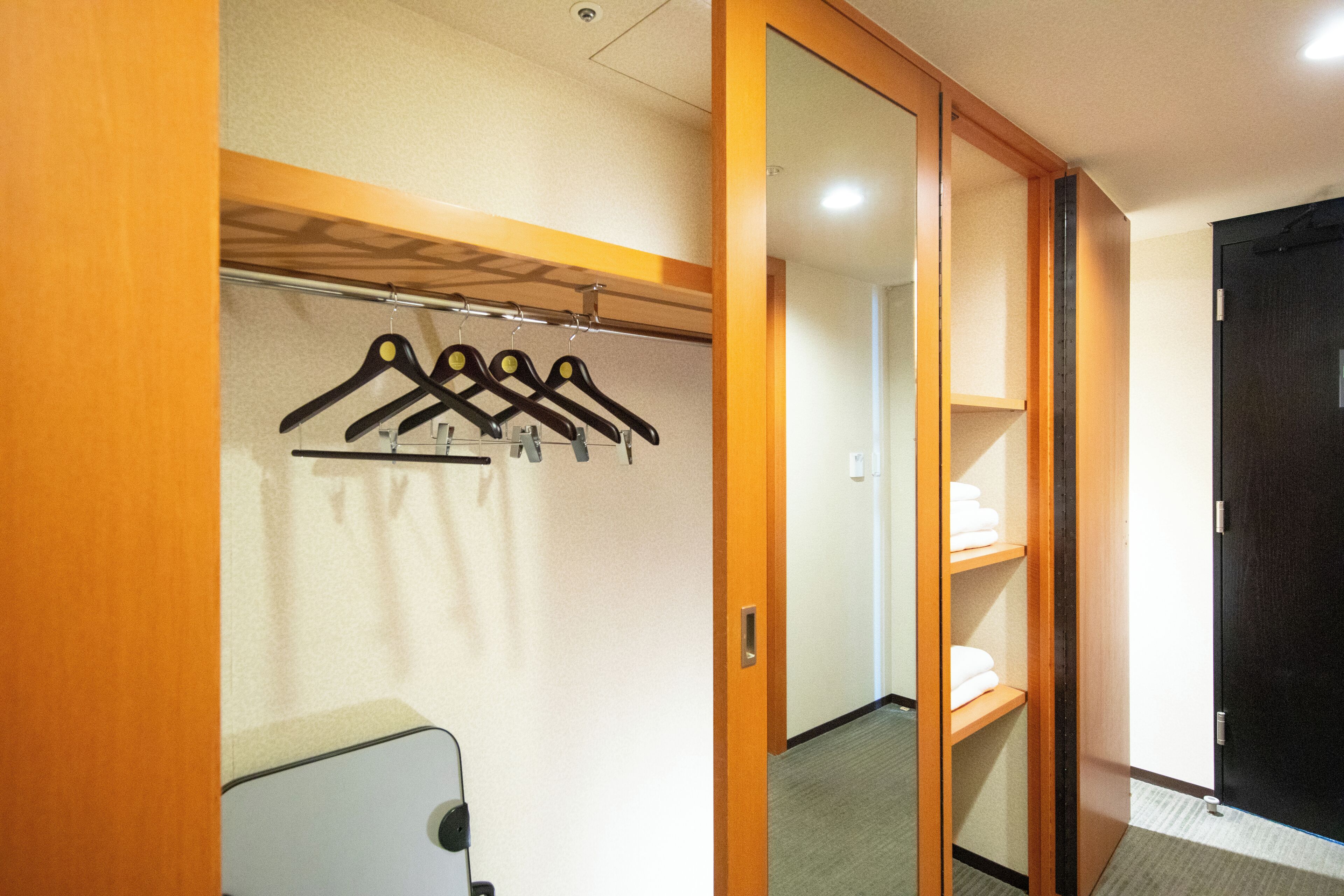 A hotel room with a wooden wardrobe, a mirror, a coat rack with hangers, and a shelf with folded towels.