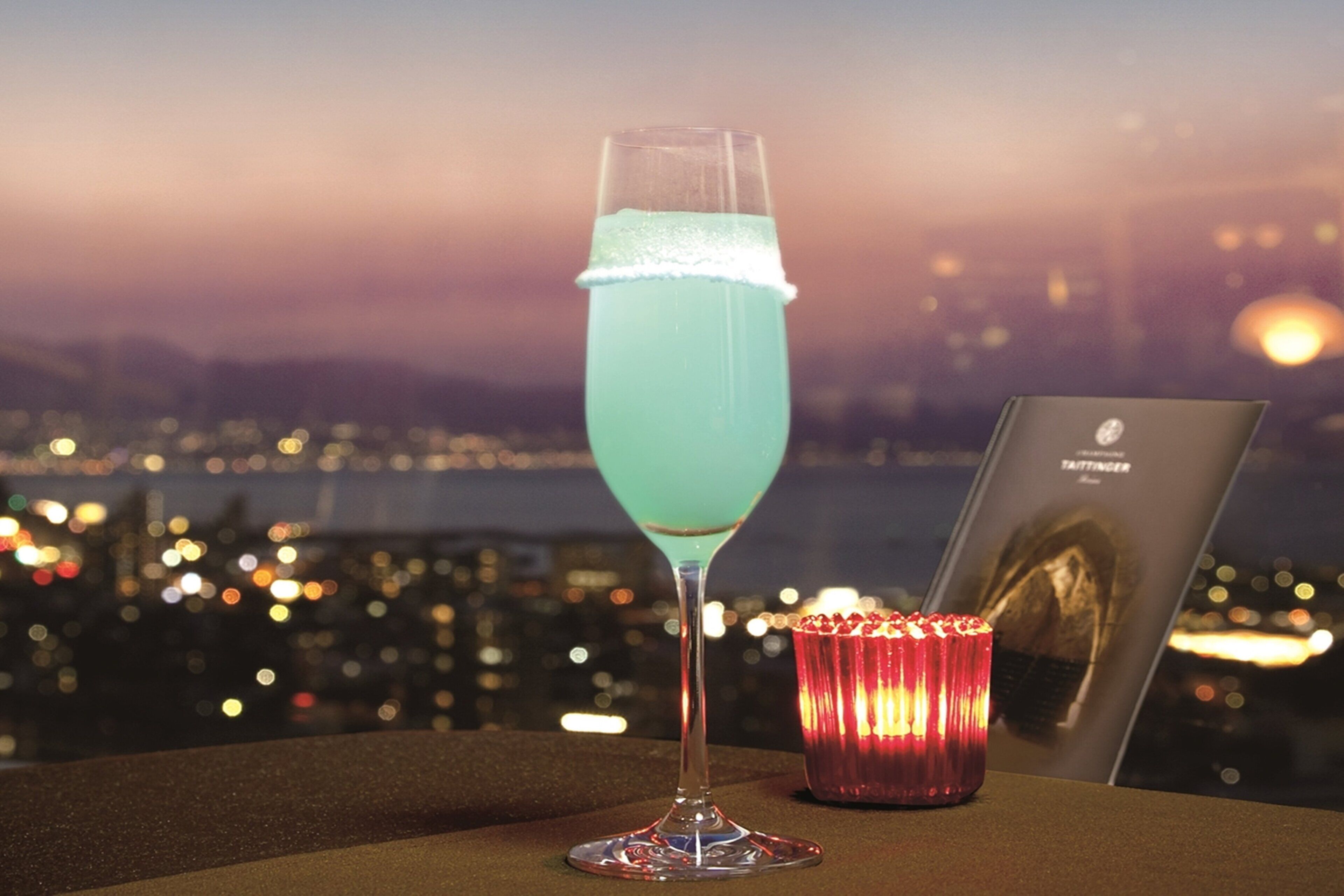 A cocktail glass with a blue liquid, a lit candle, and a framed picture on a table with a cityscape view in the background.