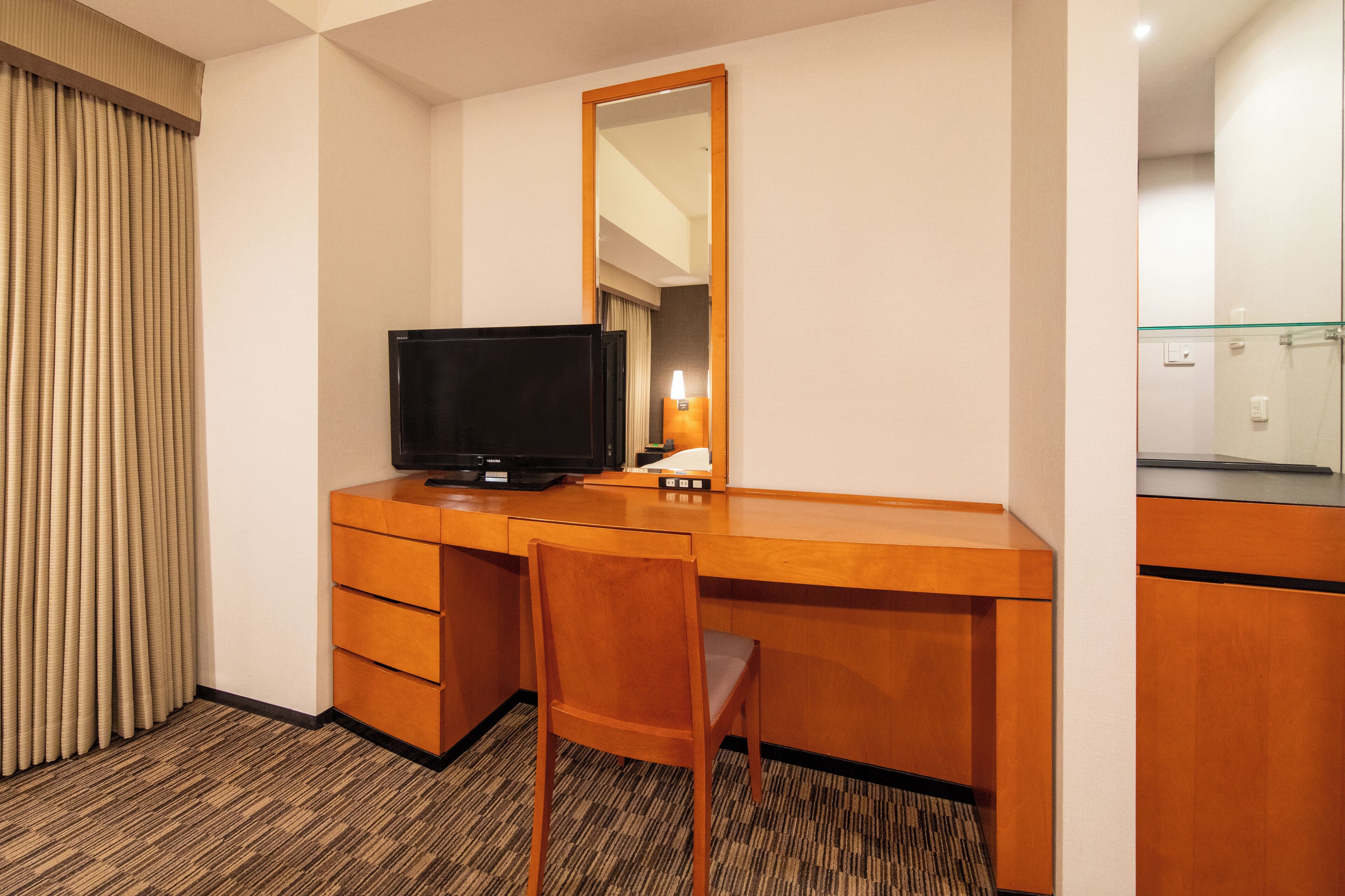 A hotel room with a wooden desk, a television, a mirror, and a glass shower enclosure.