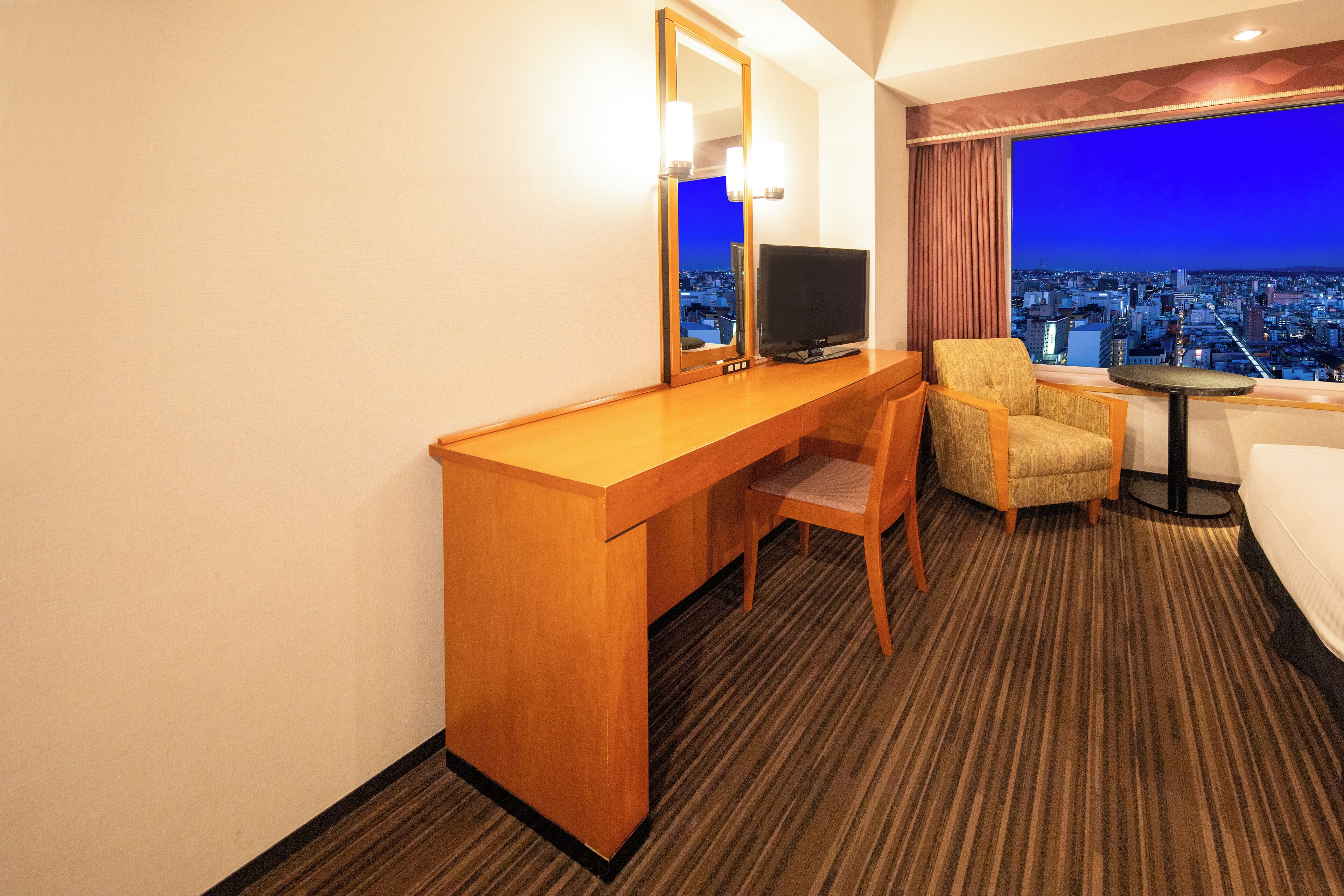 A hotel room with a wooden desk, a television, a chair, a small table, and a large window offering a city view.