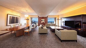 Habitación de hotel amplia con una larga mesa de comedor, asientos cómodos, un televisor de pantalla plana y un gran ventanal con vistas panorámicas.