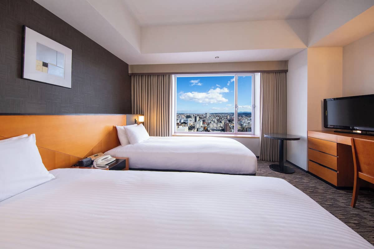 Habitació d'hotel amb dos llits, un televisor, un escriptori i una finestra gran amb vista a la ciutat.