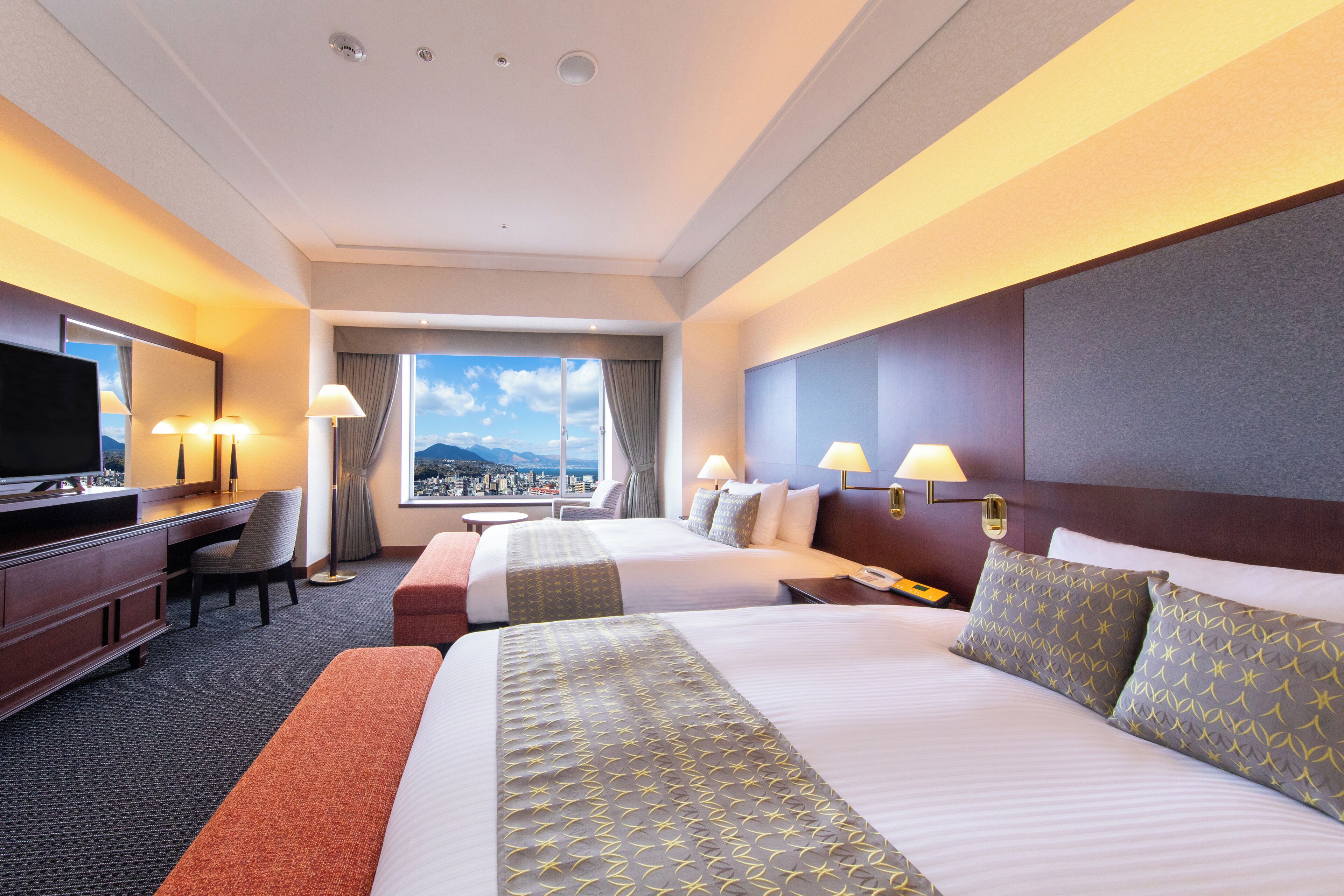 A hotel room with two beds, a desk, a TV, and a city view.