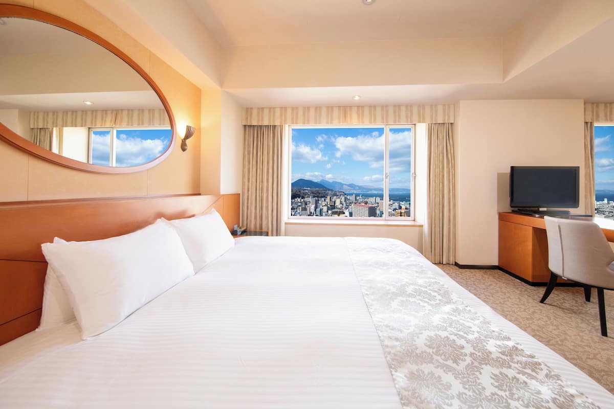 Habitació d'hotel amb un llit gran, una taula amb cadira, televisió i vista a un paisatge urbà a través de grans finestres.