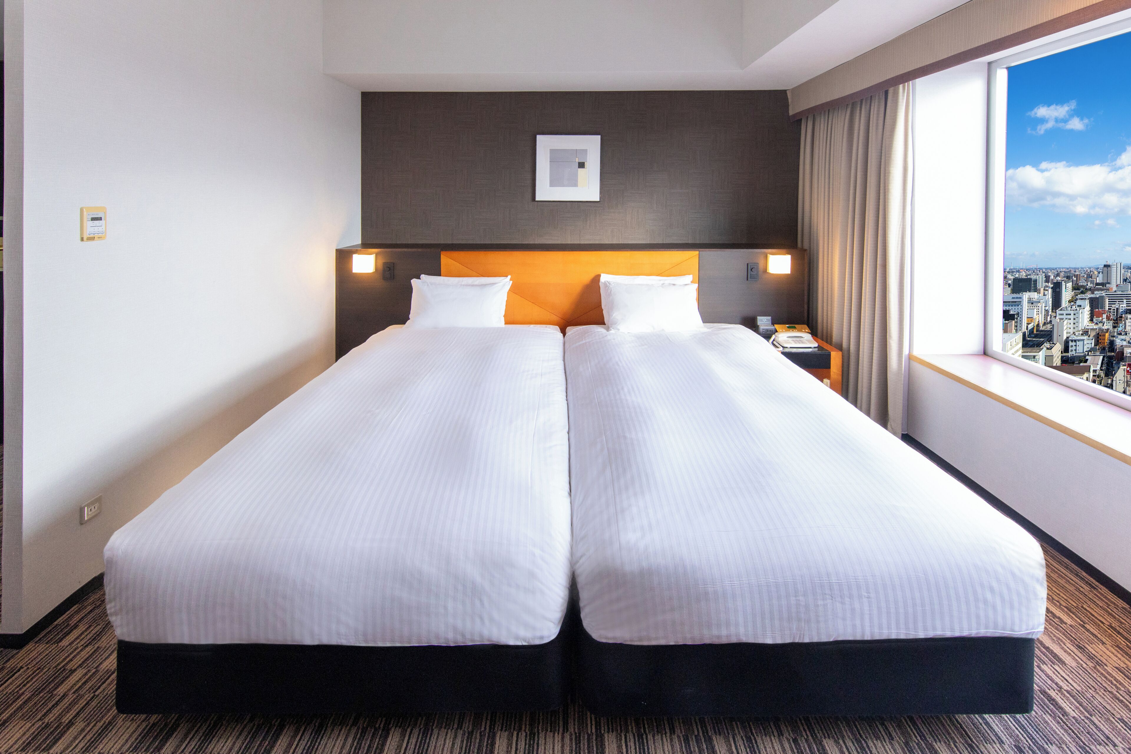 A hotel room with two single beds, a city view, a headboard with a light fixture, and a nightstand with a phone and lamp.