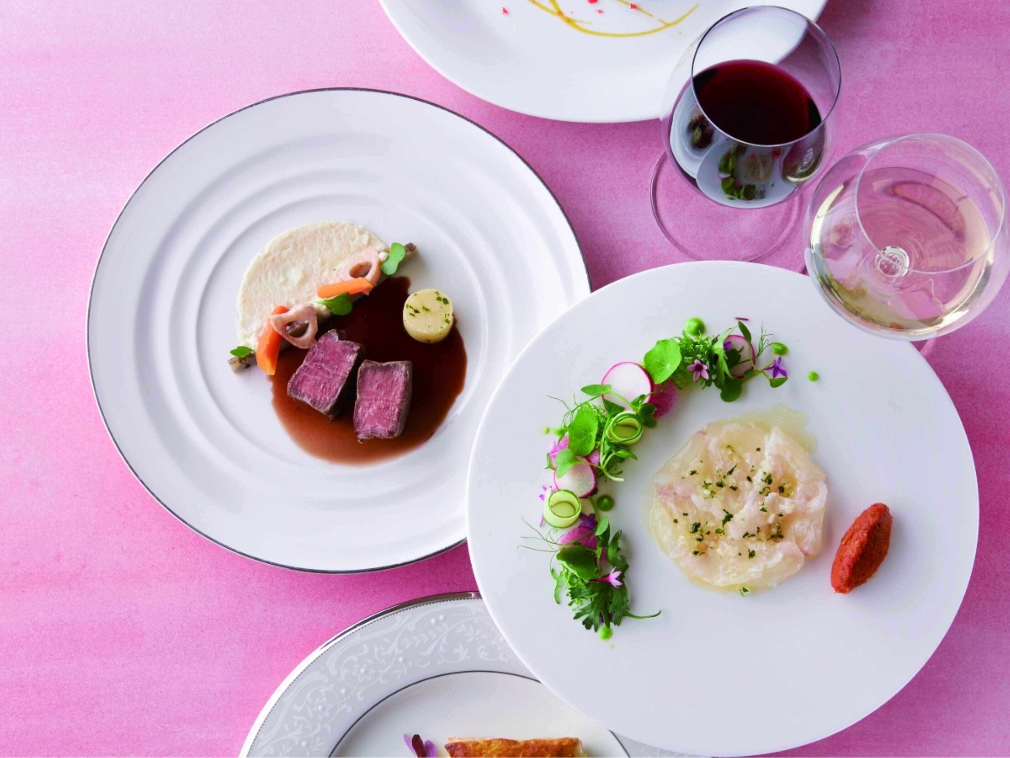 A table with three plates of food, two glasses of red wine, and a pink tablecloth.