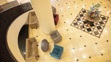 Una hall con una scalinata a forma di curva, un'area salotto centrale con divani e poltrone, una pianta decorativa e un pavimento a motivi.