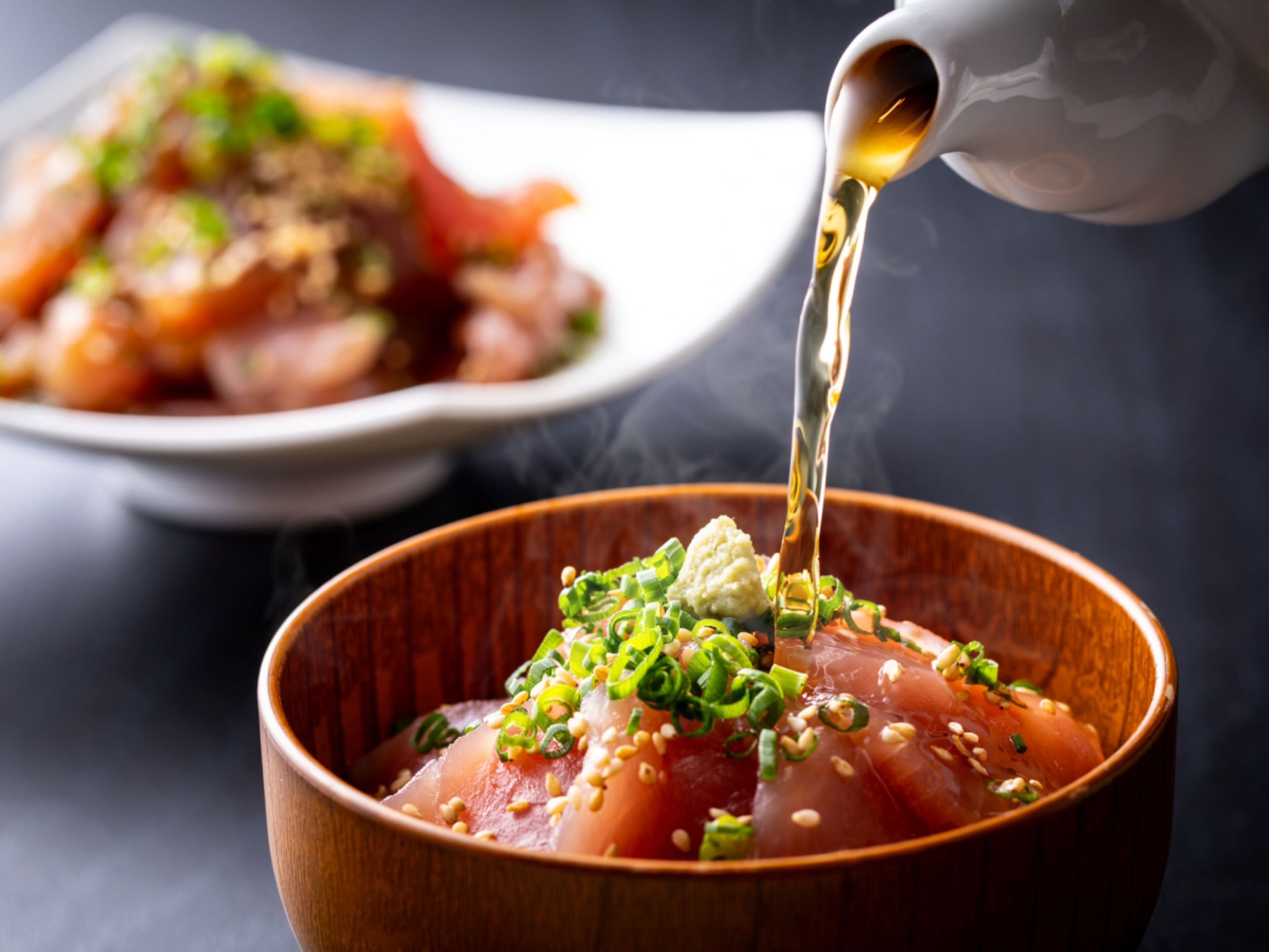 A wooden bowl with a raw fish dish garnished with green onions and sesame seeds, with a sauce being poured over it.
