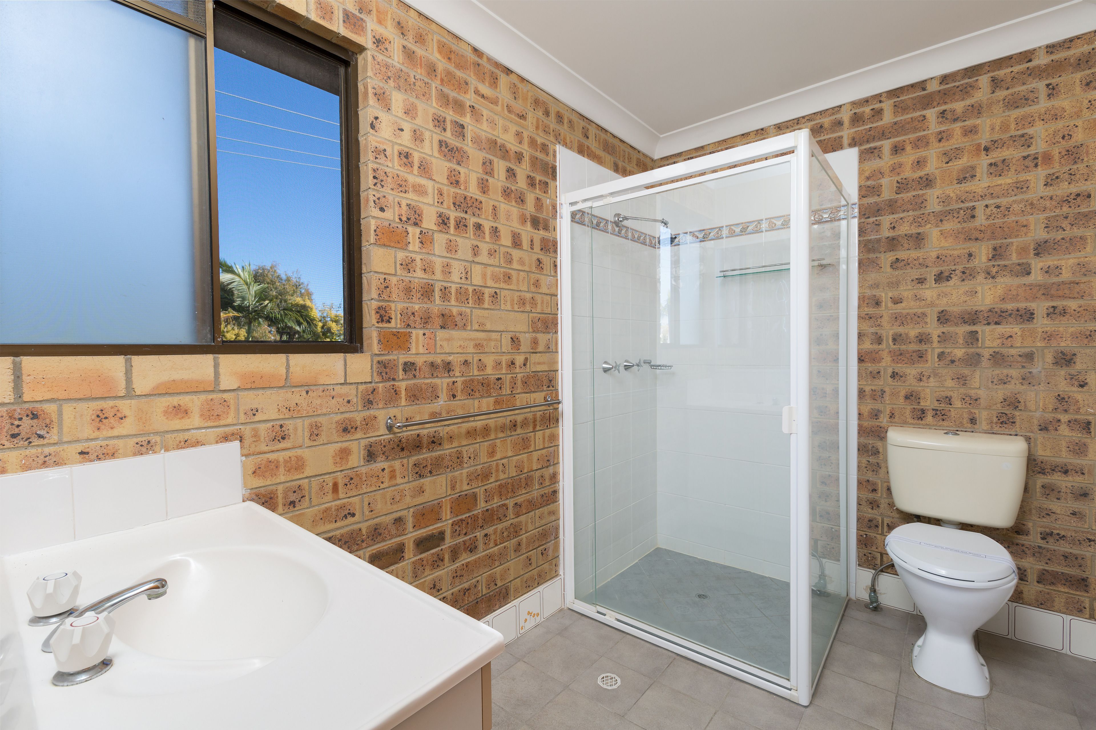 A bathroom with a white sink, a glass-enclosed shower, and a toilet against a brick wall.