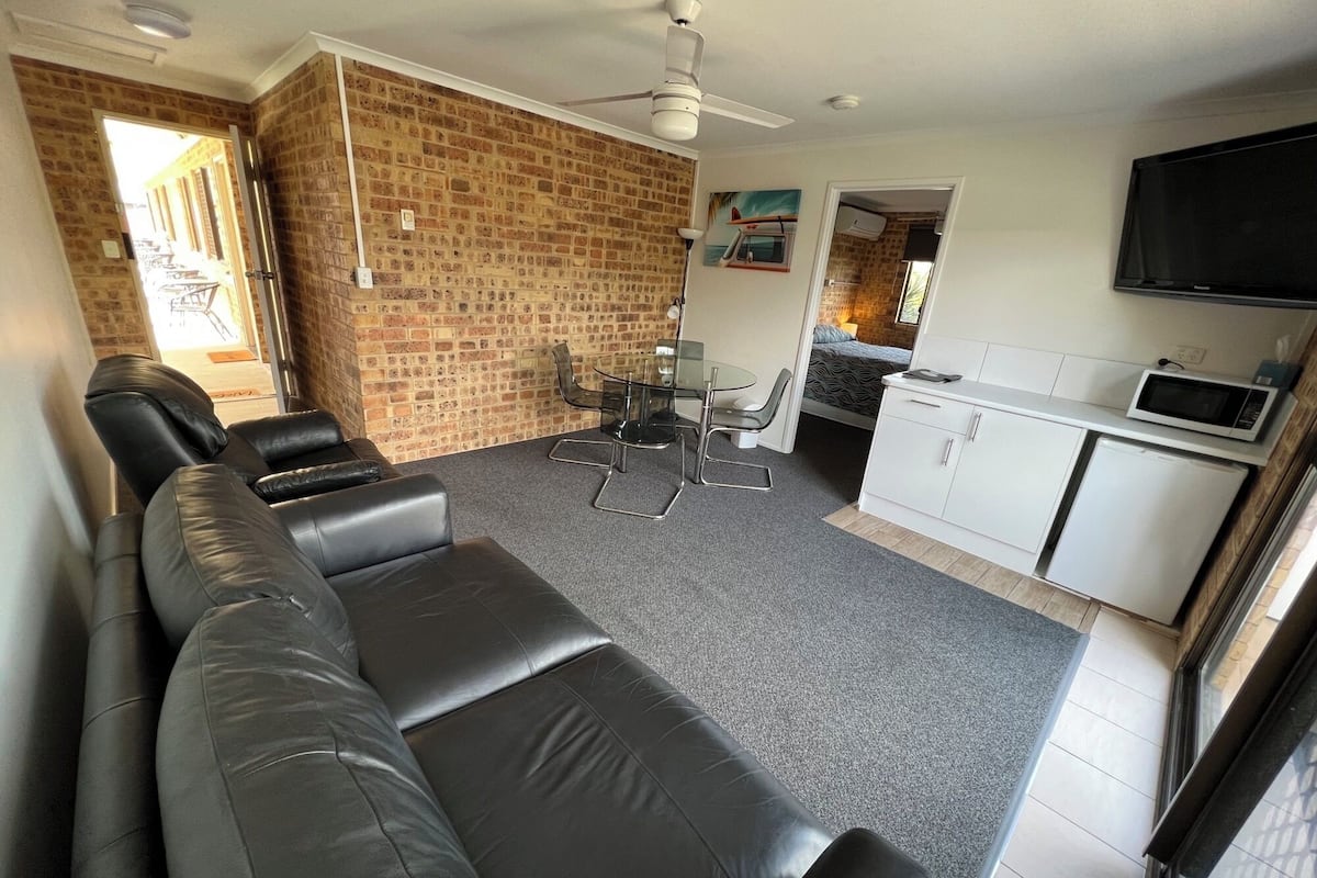 A living room with a black leather sofa, a glass dining table with chairs, a kitchen area with a microwave, and a brick wall.