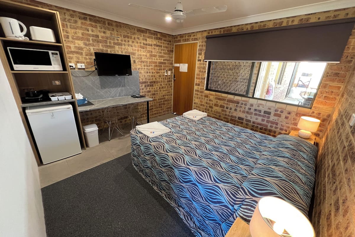 A compact hotel room with a kitchenette, a bed with patterned bedding, a desk with a flat-screen TV, and a window with a view.