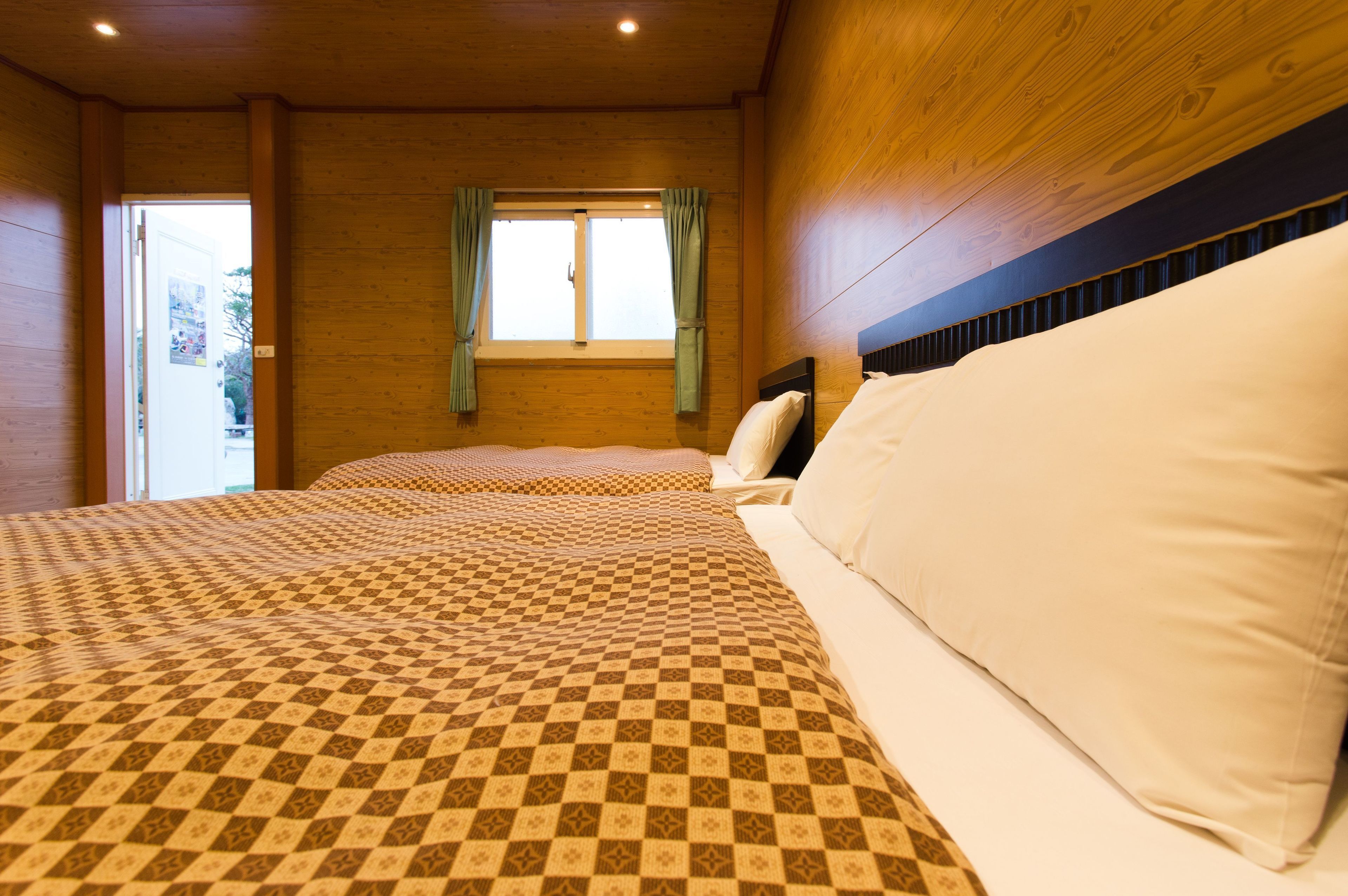 A room with two beds, wooden walls, a window with curtains, and a view of the outside.