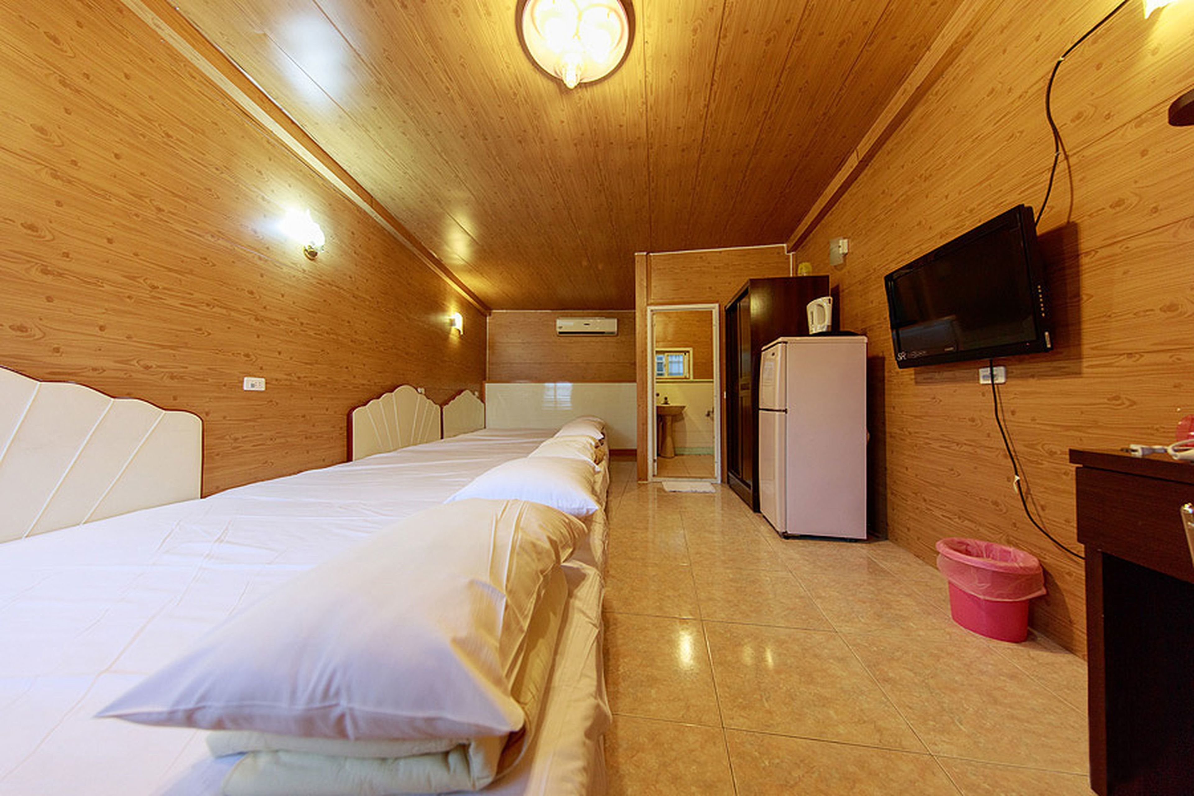 A hotel room with wooden walls, a bed with white bedding, a television mounted on the wall, and a small refrigerator.
