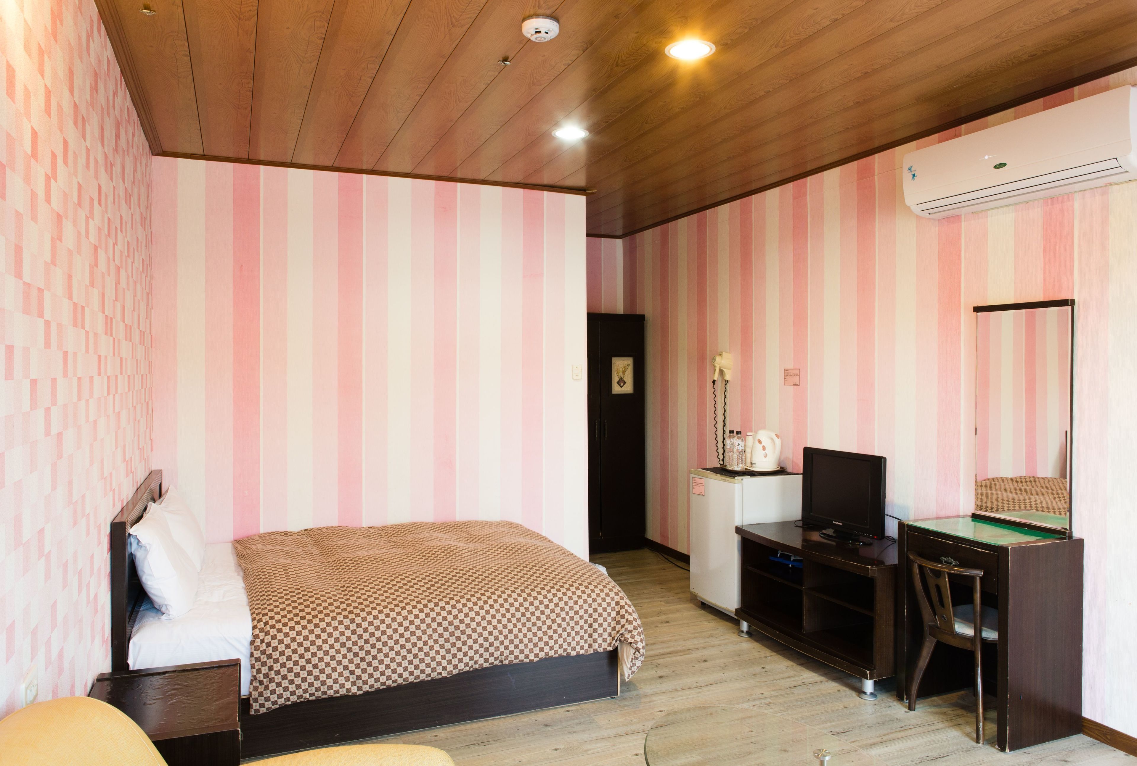 A bedroom with a bed, a desk, a chair, a television, and an air conditioning unit.