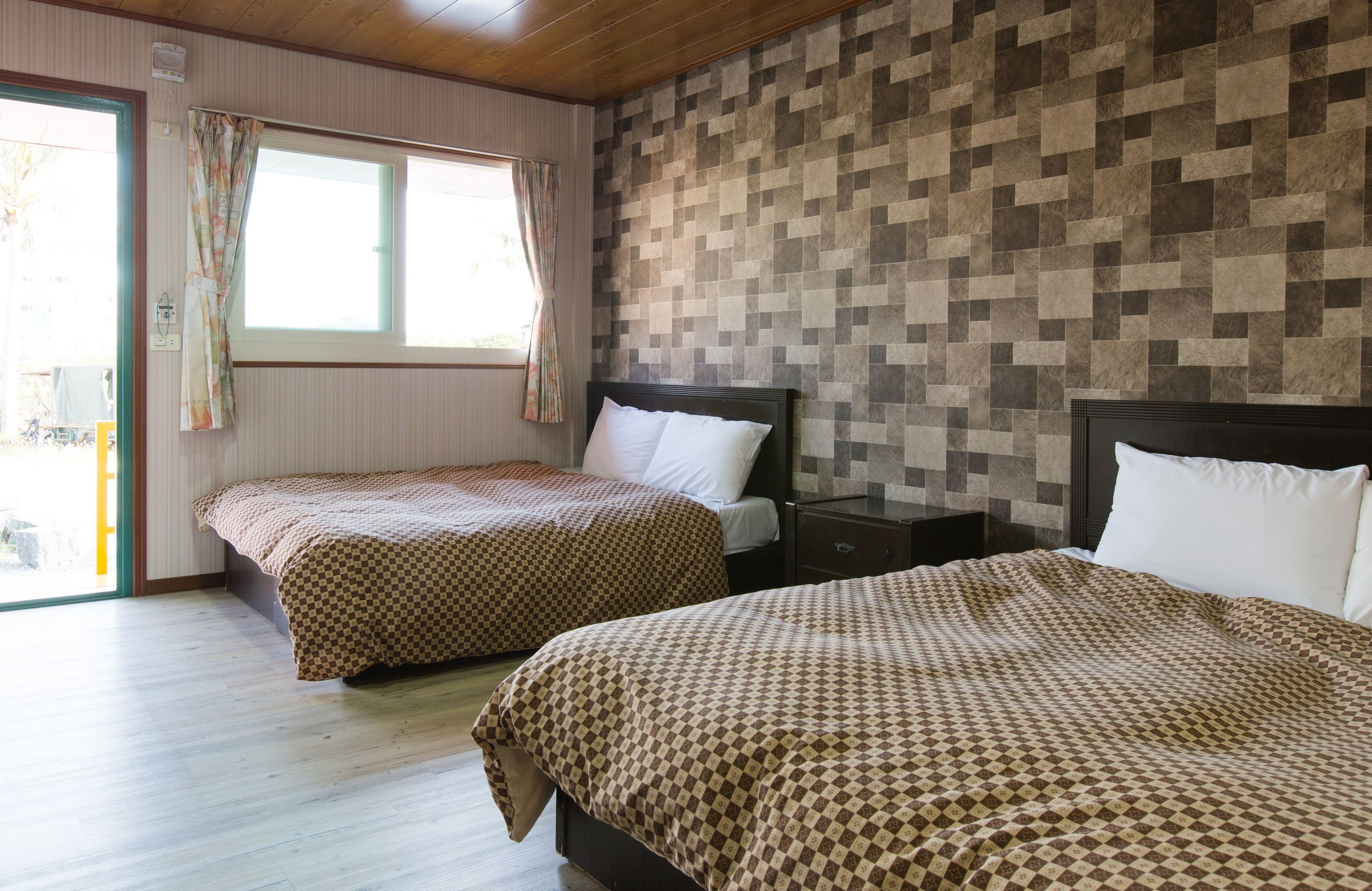 A room with two beds, a window with curtains, a checkered wall pattern, and a wooden floor.