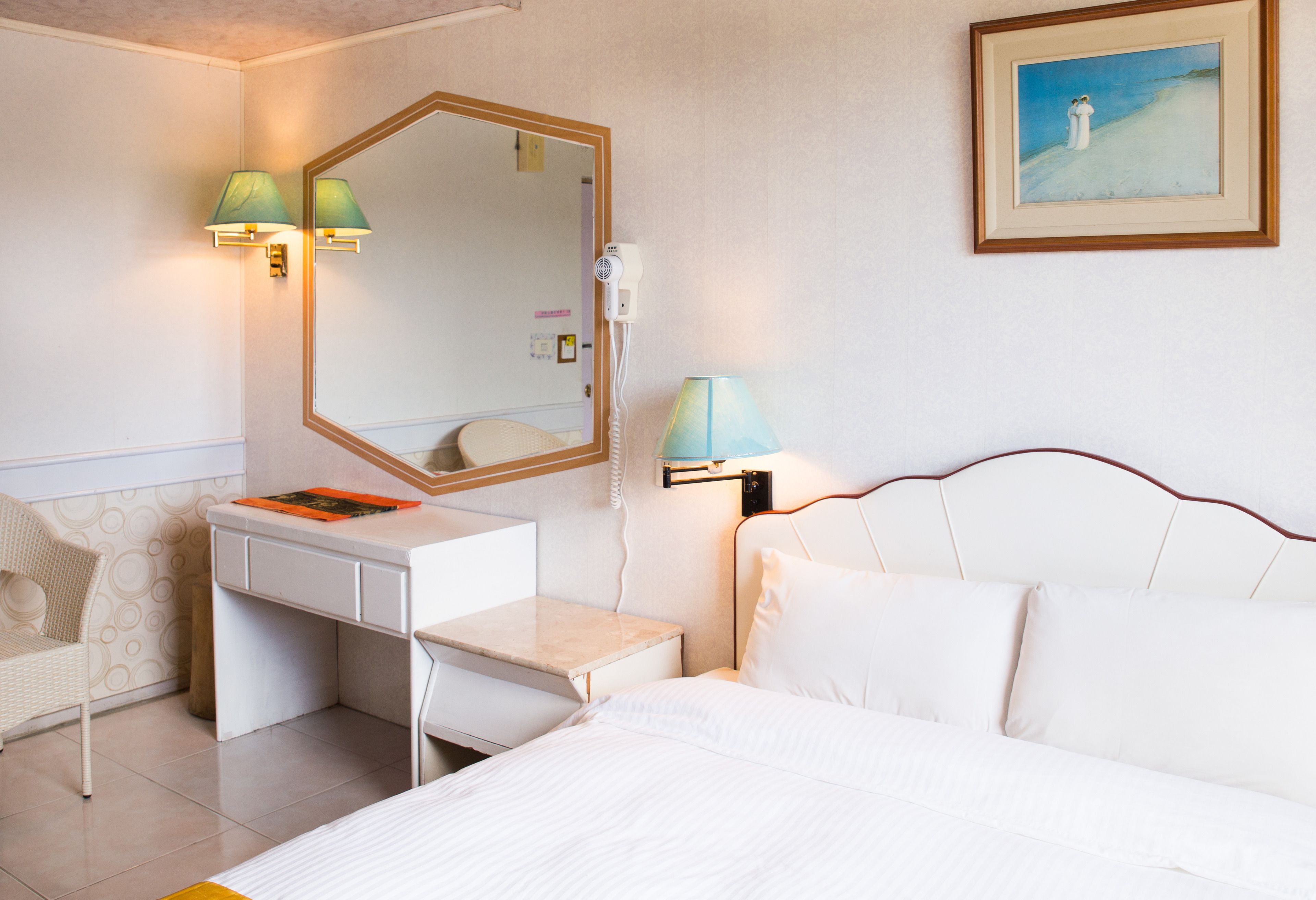 A hotel room with a bed, a desk with a chair, a lamp, a mirror, and a painting on the wall.
