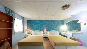 A hotel room with two single beds, a window with curtains, a wall mural, a desk with a lamp, and a shelf with a bag. - Dajenshan Hotel (Hengchun)