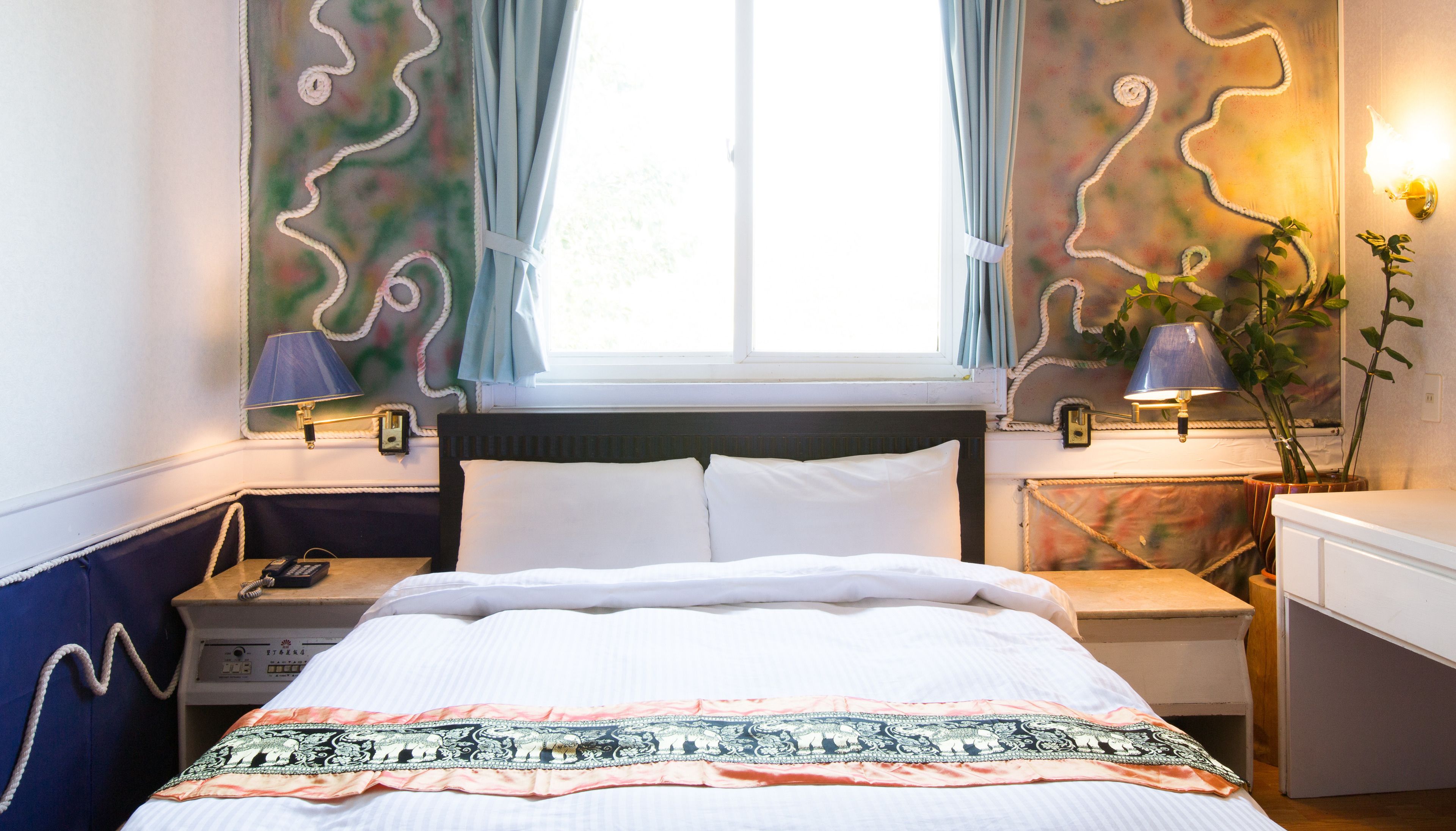A neatly made bed with a patterned bedspread, two bedside lamps, a nightstand with a phone, and a window with curtains.