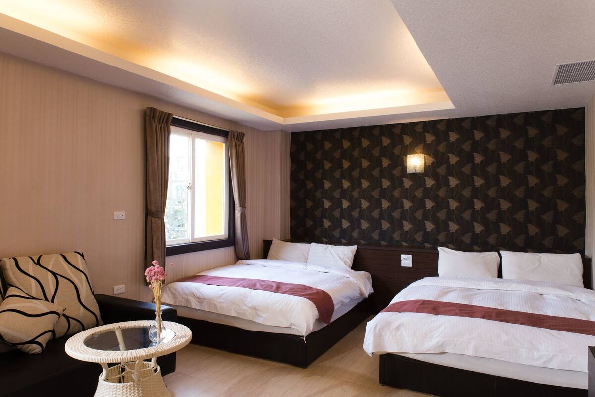 Ett hotellrum med två sängar, ett litet bord med en vas, en soffa och ett fönster med gardiner.