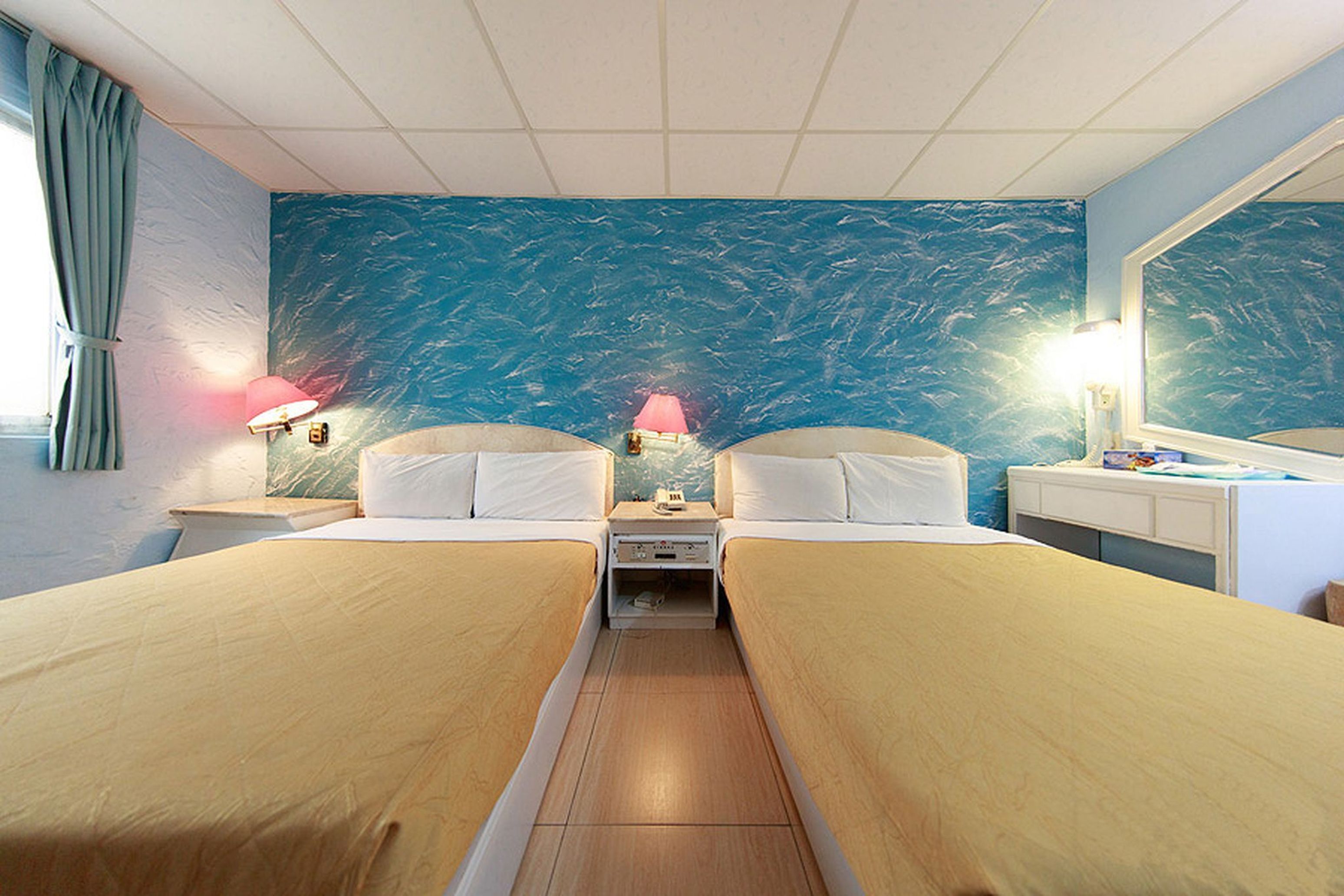 A hotel room with two single beds, a nightstand with a lamp, a dresser, and a large mirror.
