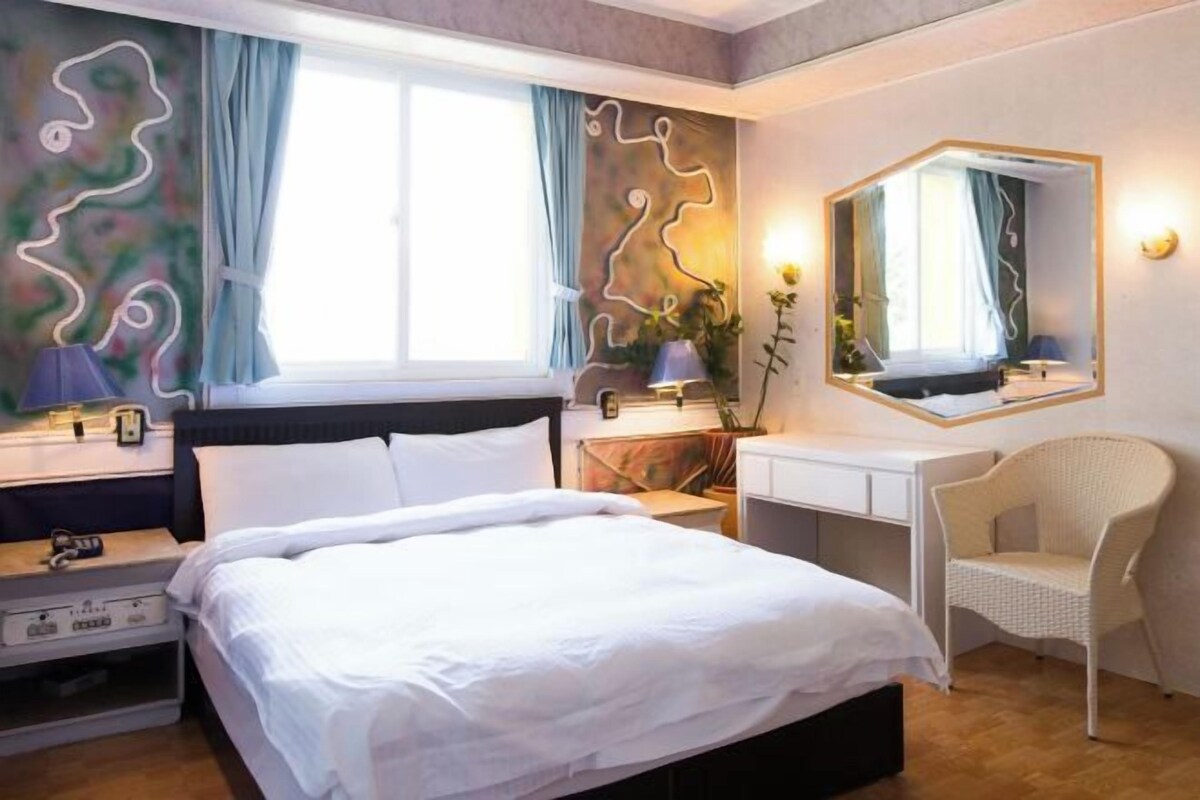 A hotel room with a bed, a desk, a chair, a mirror, and a window with curtains.