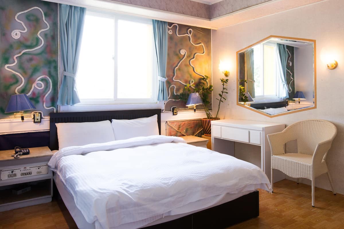 Ett hotellrum med en säng, ett skrivbord, en stol, en spegel och ett fönster med gardiner.