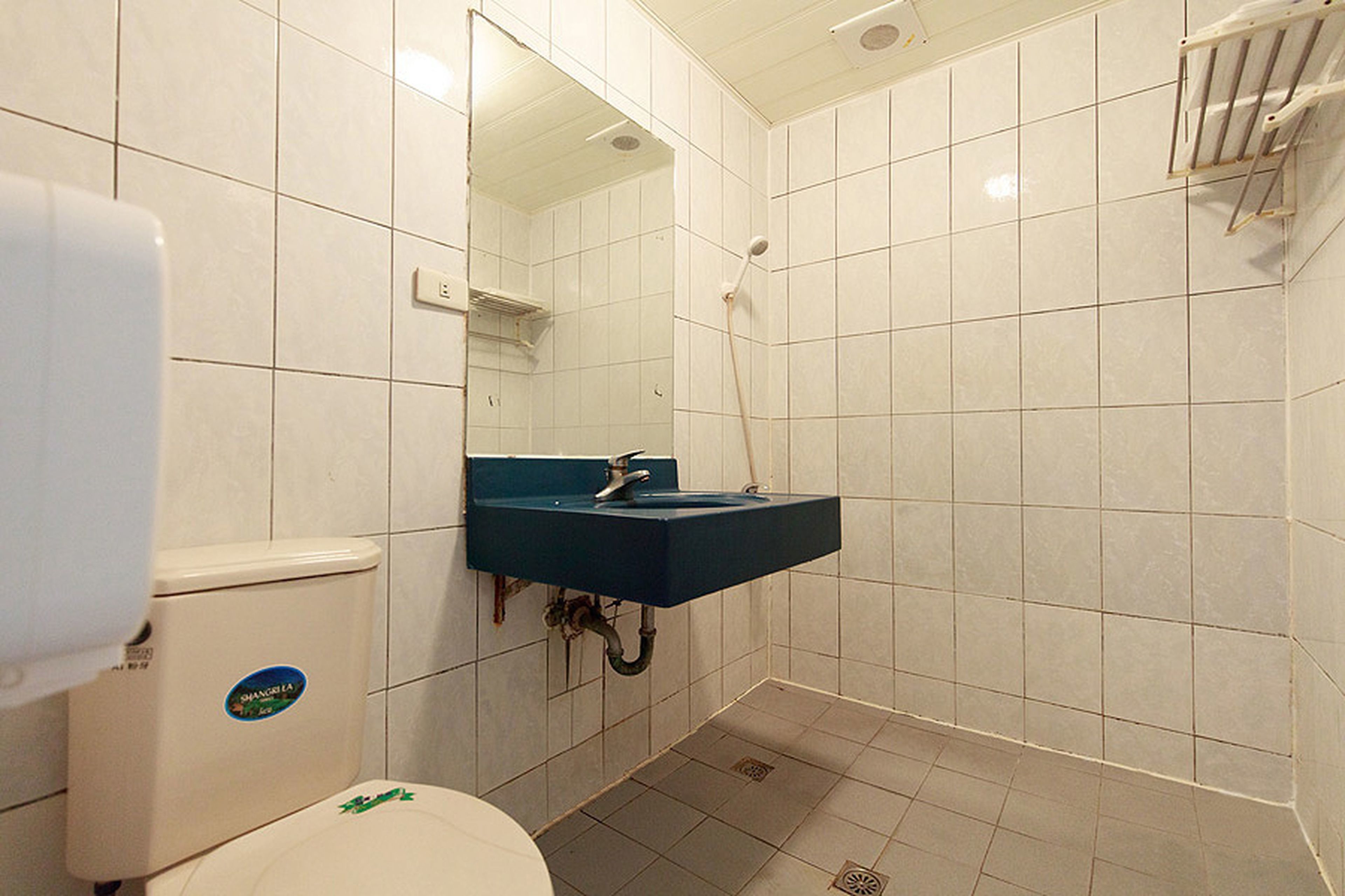 Un baño pequeño con paredes de azulejos blancos, un inodoro blanco, un lavamanos azul y una zona de ducha.
