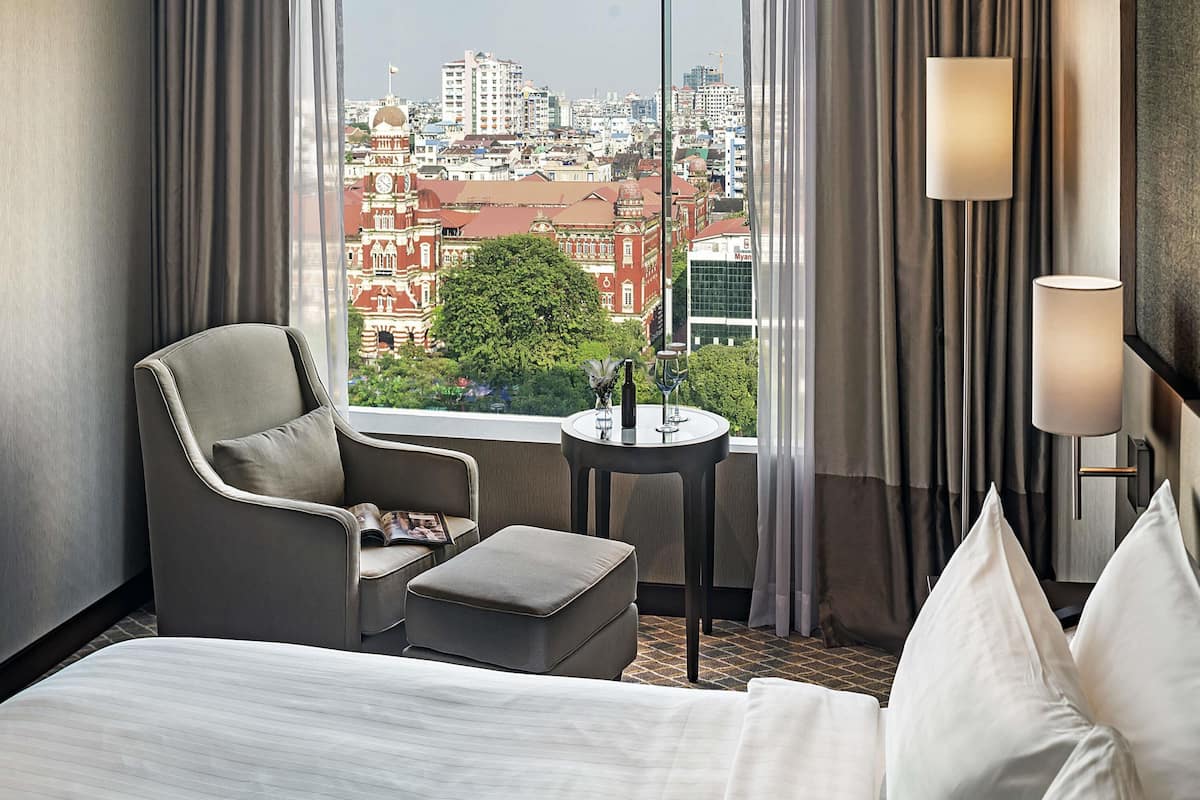 Ein Hotelzimmer mit Bett, einem Sessel, einem kleinen Tisch mit Flasche und Gläsern sowie Blick auf eine Stadtkulisse durch große Fenster.
