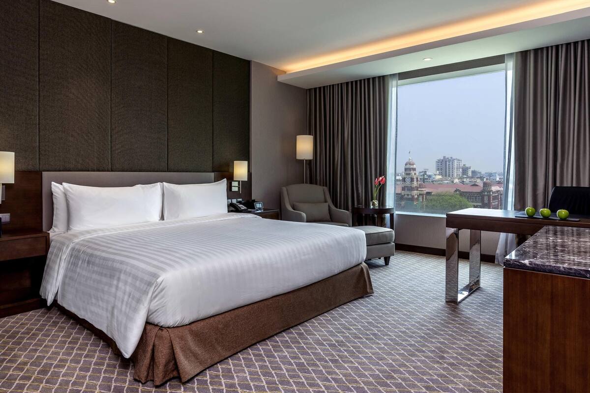 Ein Hotelzimmer mit einem großen Bett, einem Schreibtisch, einem Stuhl, einer Lampe und Blick auf die Stadt.